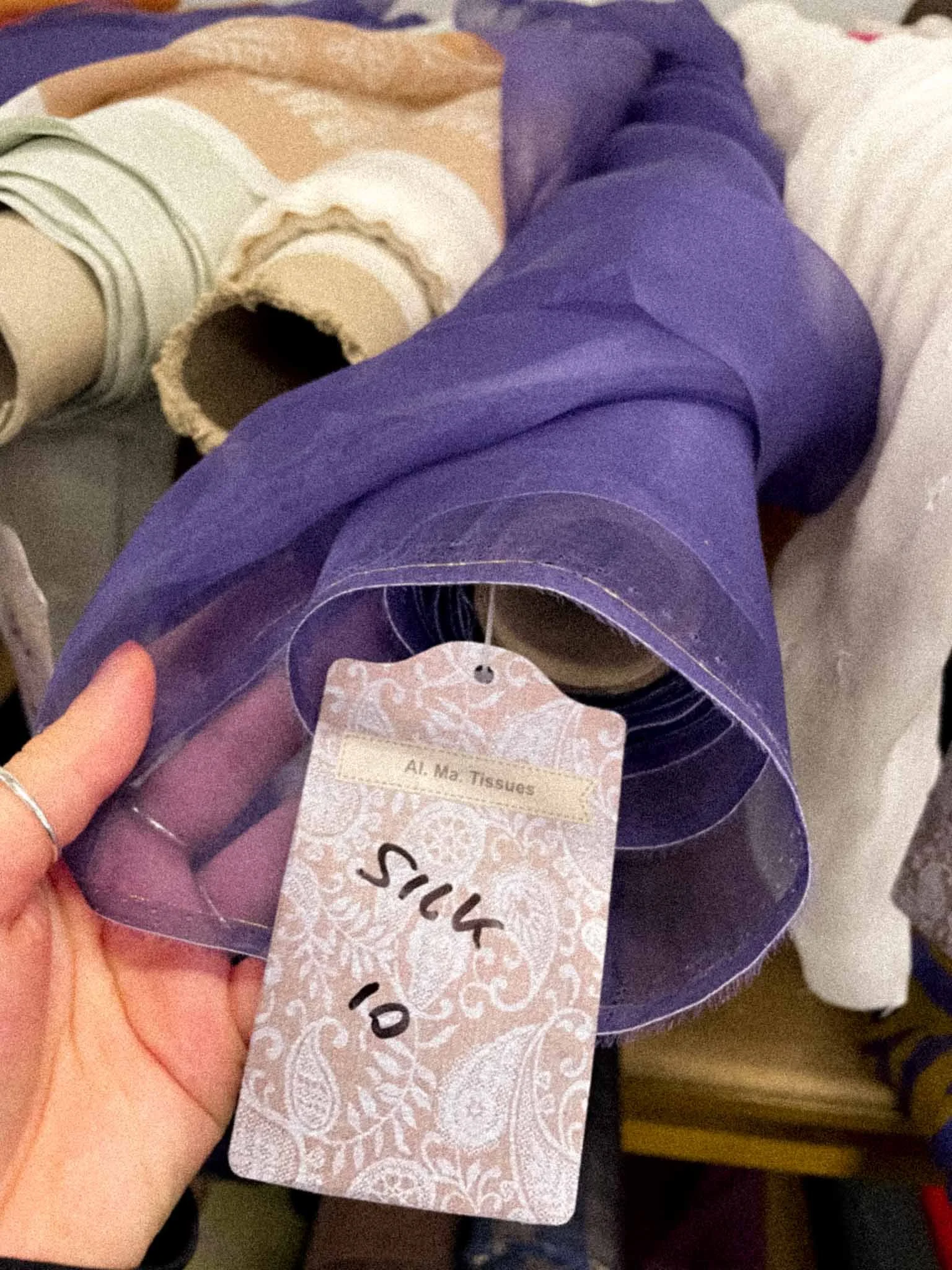 A hand holding a small package of tissues labeled 'Al.Ma. Tissues,' with various partially visible fabric items in the background.