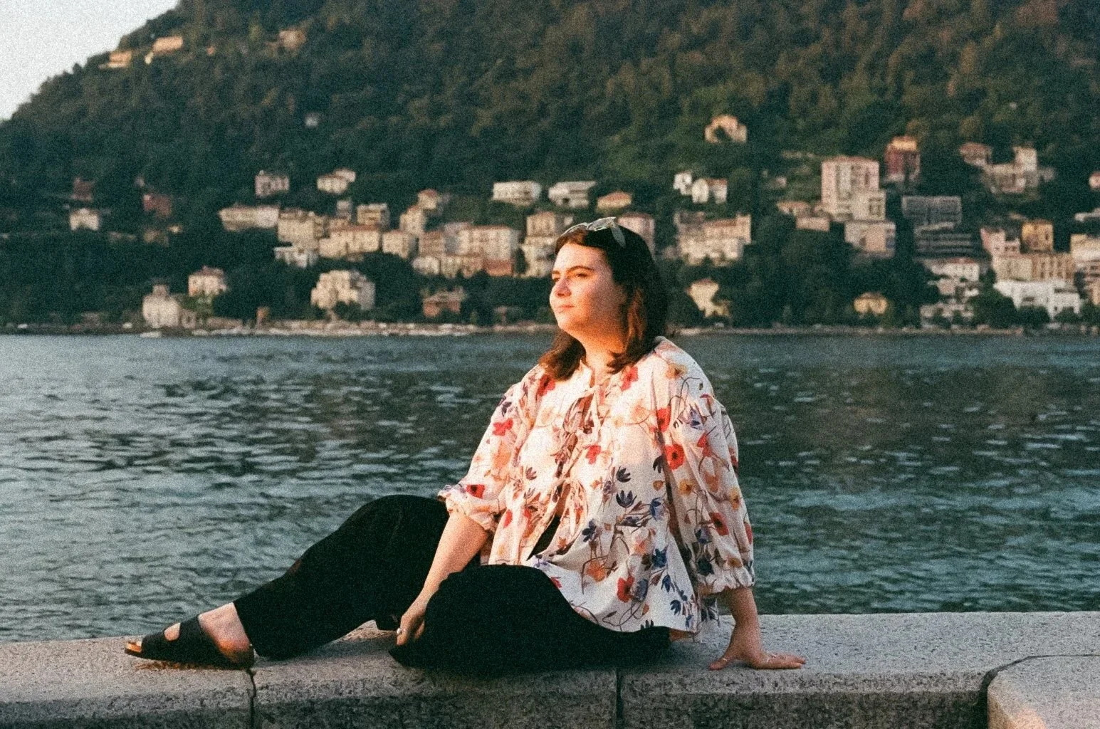 A woman sits on a stone ledge by the water with a city and hillside in the background, her eyes closed, enjoying the sunset.