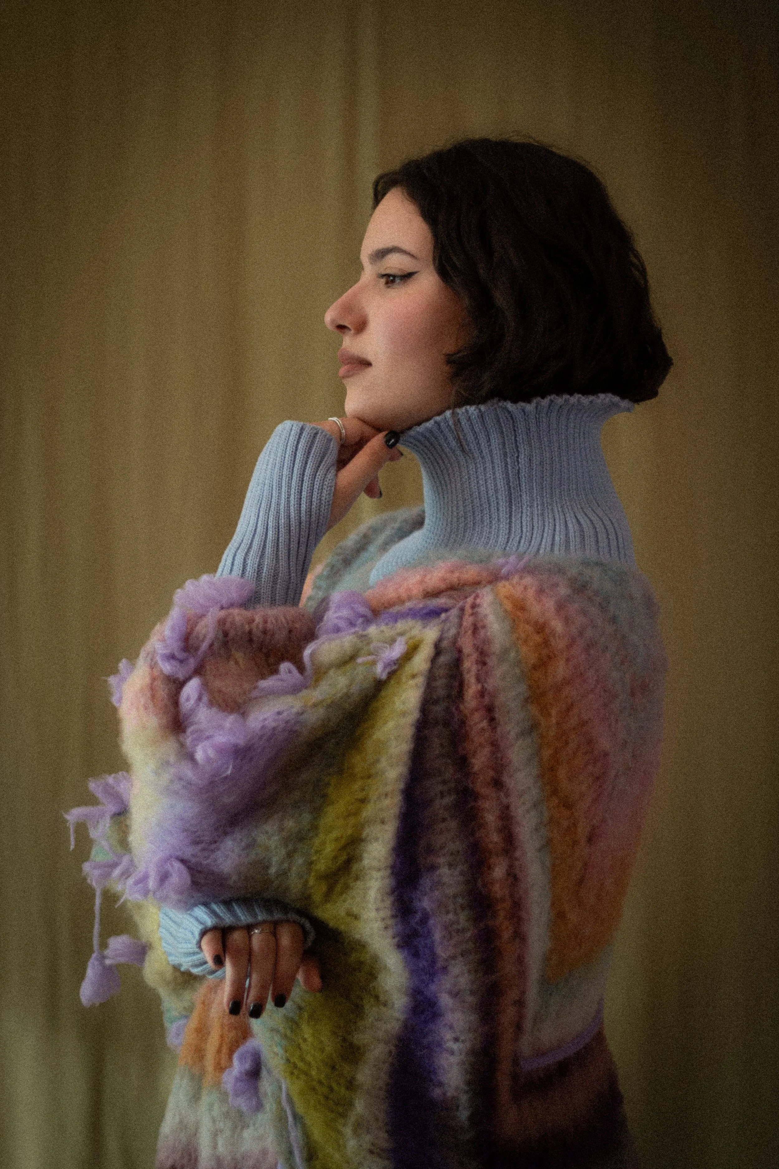 A woman with short black hair and light makeup wearing a pastel-colored turtleneck sweater and a multicolored, textured shawl with pom-poms on the edges, standing against a plain, beige background.