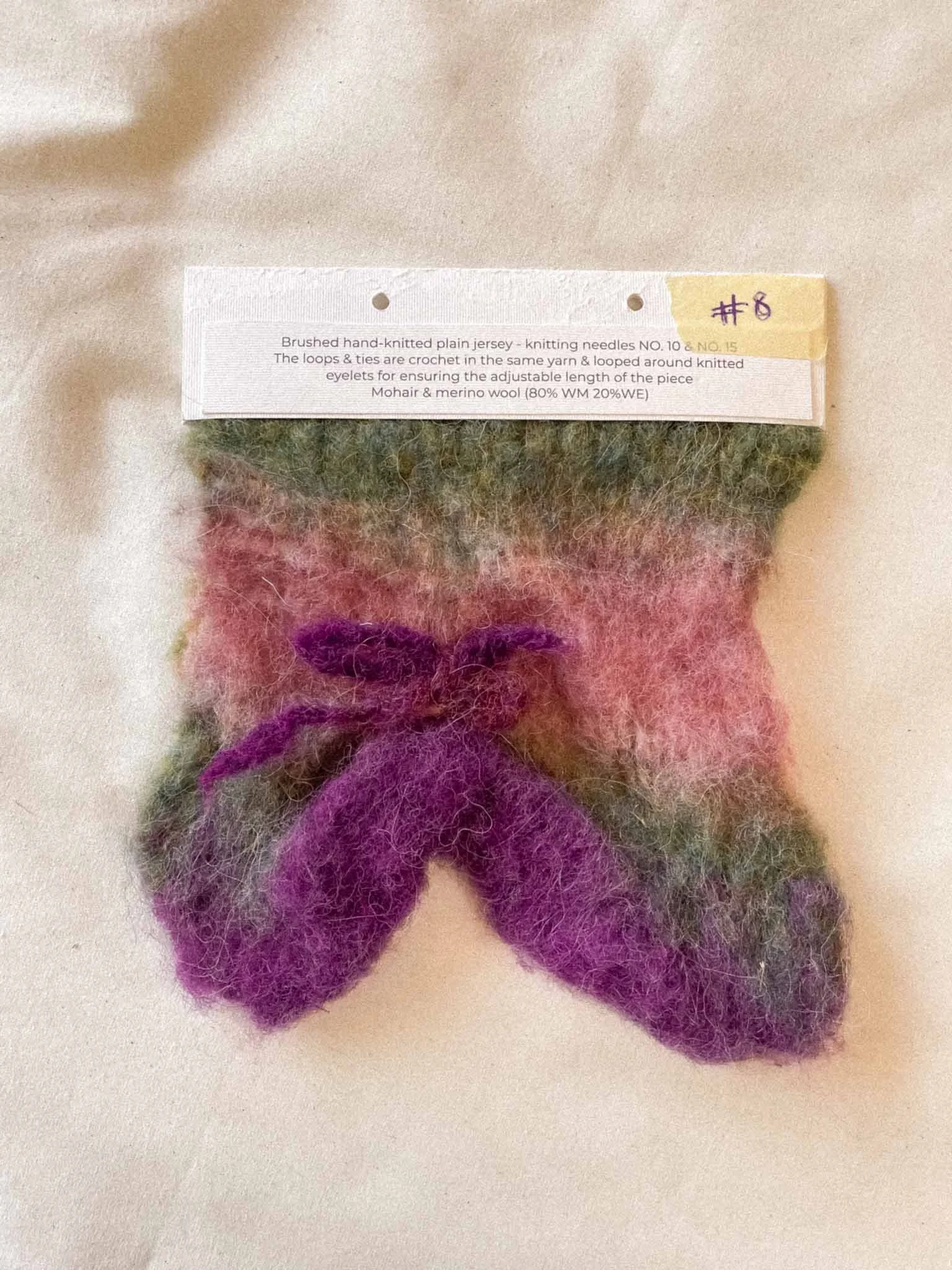 Colorful felted wool flower with multiple purple, pink, green, and red petals on a white background.