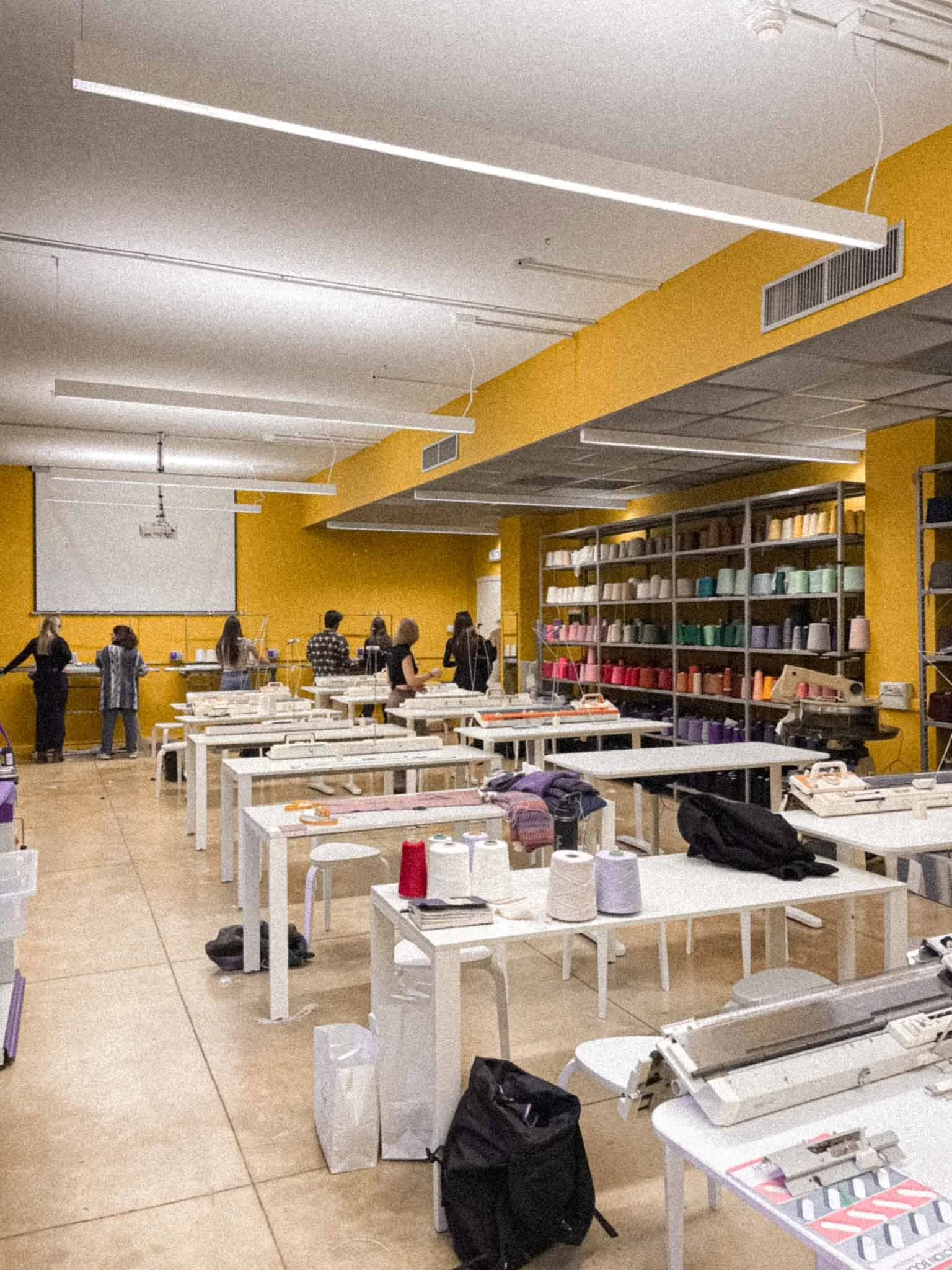 A classroom or workshop space with white tables, colorful spools of thread on shelves, and people working or shopping.