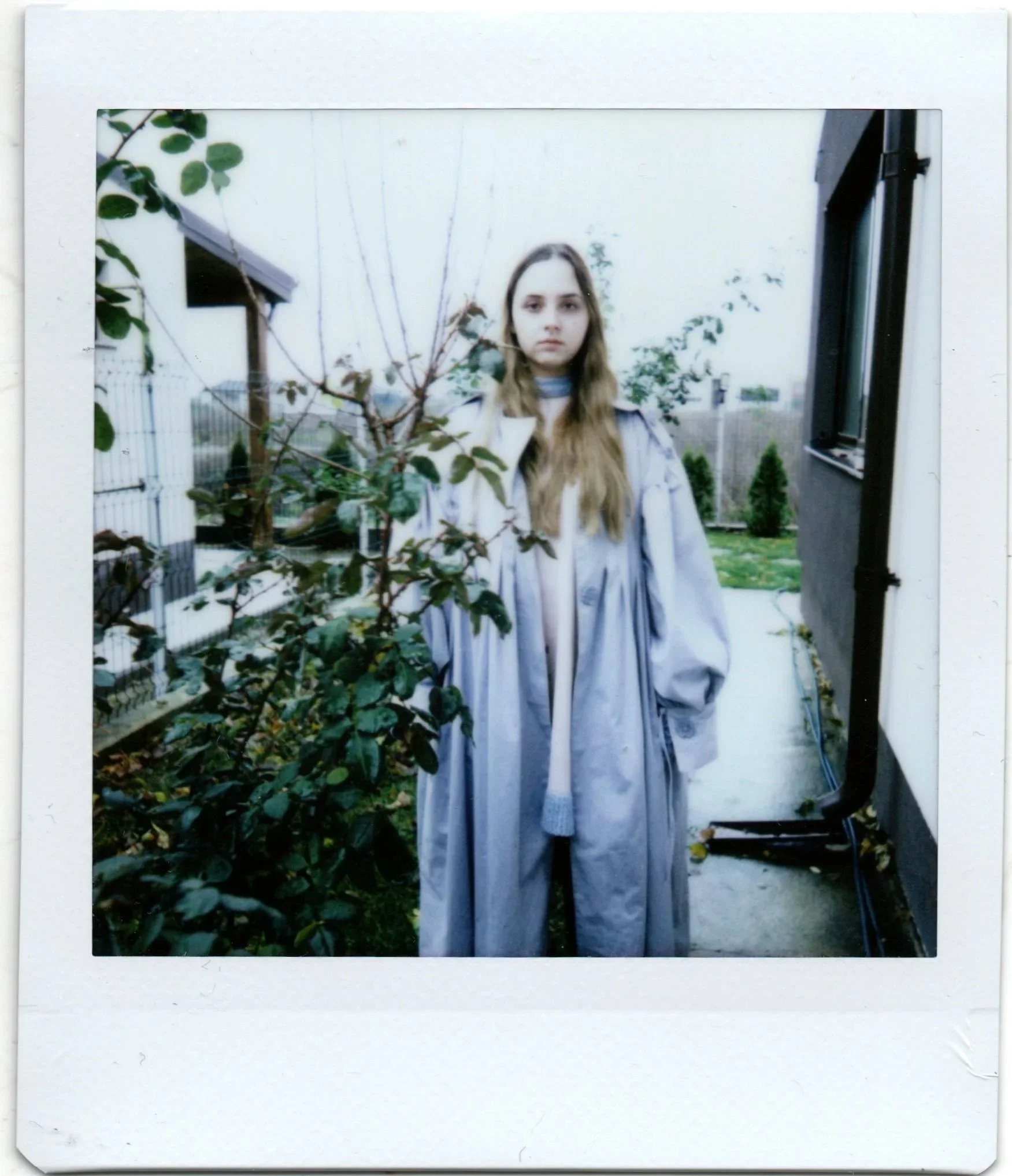 A young woman with long blonde hair wearing a light-colored jacket standing outside near a window, with a small tree and plants in the background, in an overcast day.