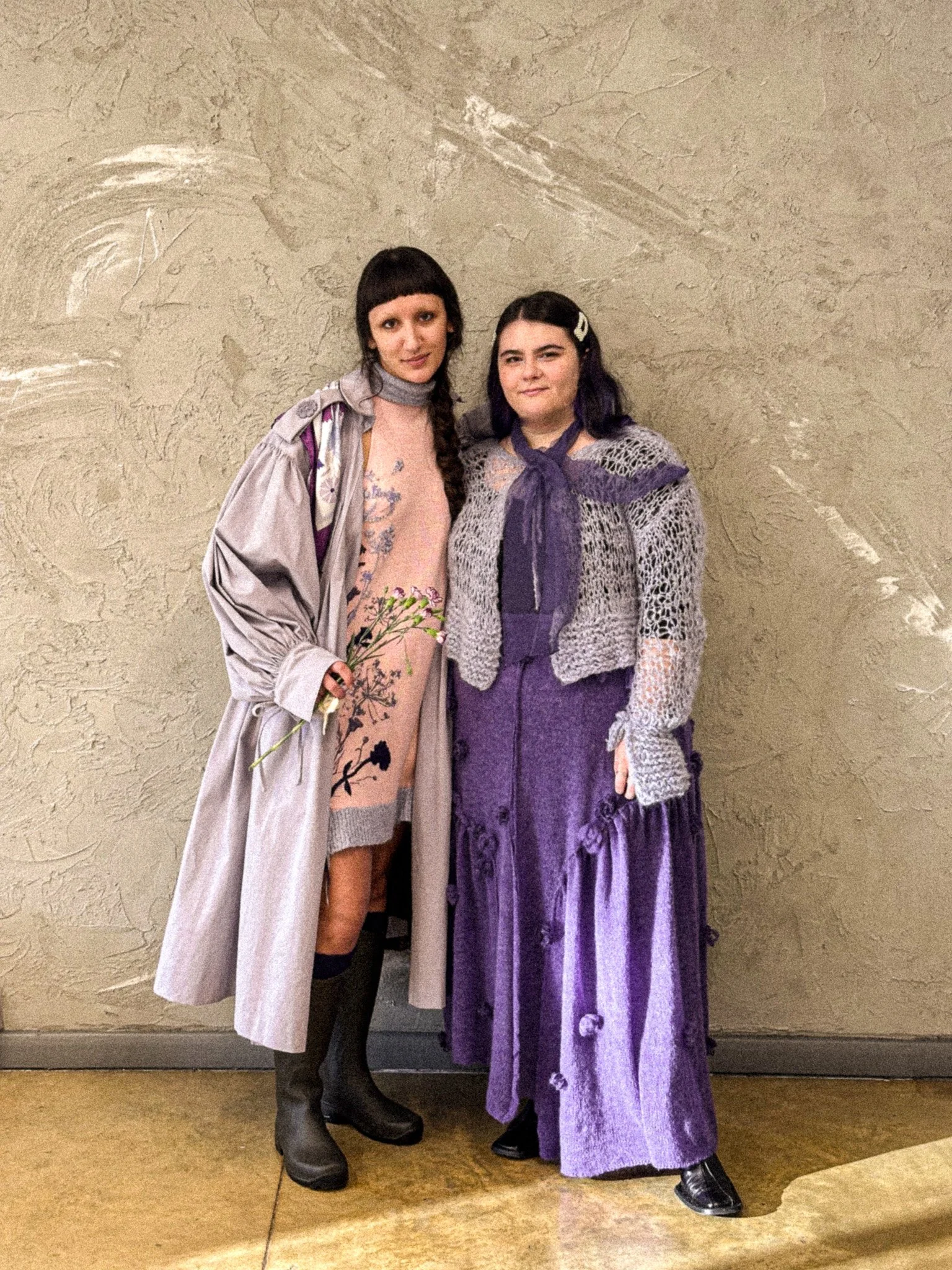 Two young women standing against a beige, textured wall, dressed in vintage or costume clothing, with one holding a small bouquet of flowers.