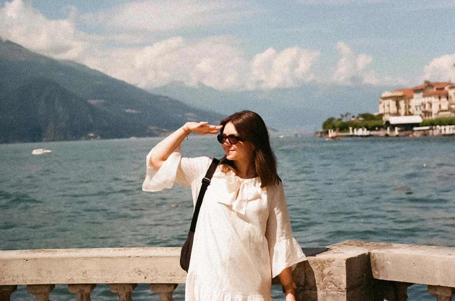 A woman wearing sunglasses and a cream-colored dress standing near a body of water with mountains and buildings in the background, shielding her eyes from the sun.