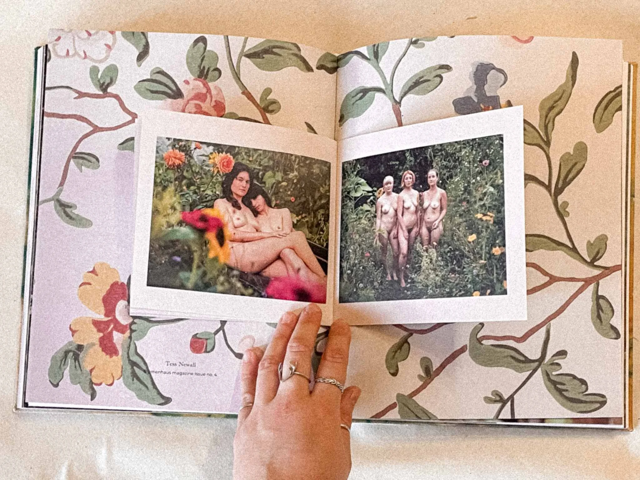 A hand holding an open magazine displaying two photographs of nude women standing in a garden, with a decorative floral border surrounding the magazine.