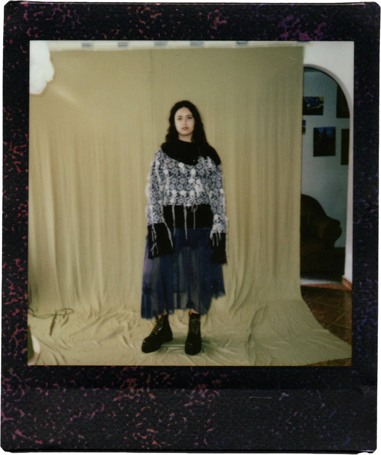 A young woman standing in front of a yellow curtain backdrop, wearing a black and white patterned sweater, a dark blue tulle skirt, and black lace-up boots with heels, in an indoor setting with paintings on the wall visible through an arched doorway.