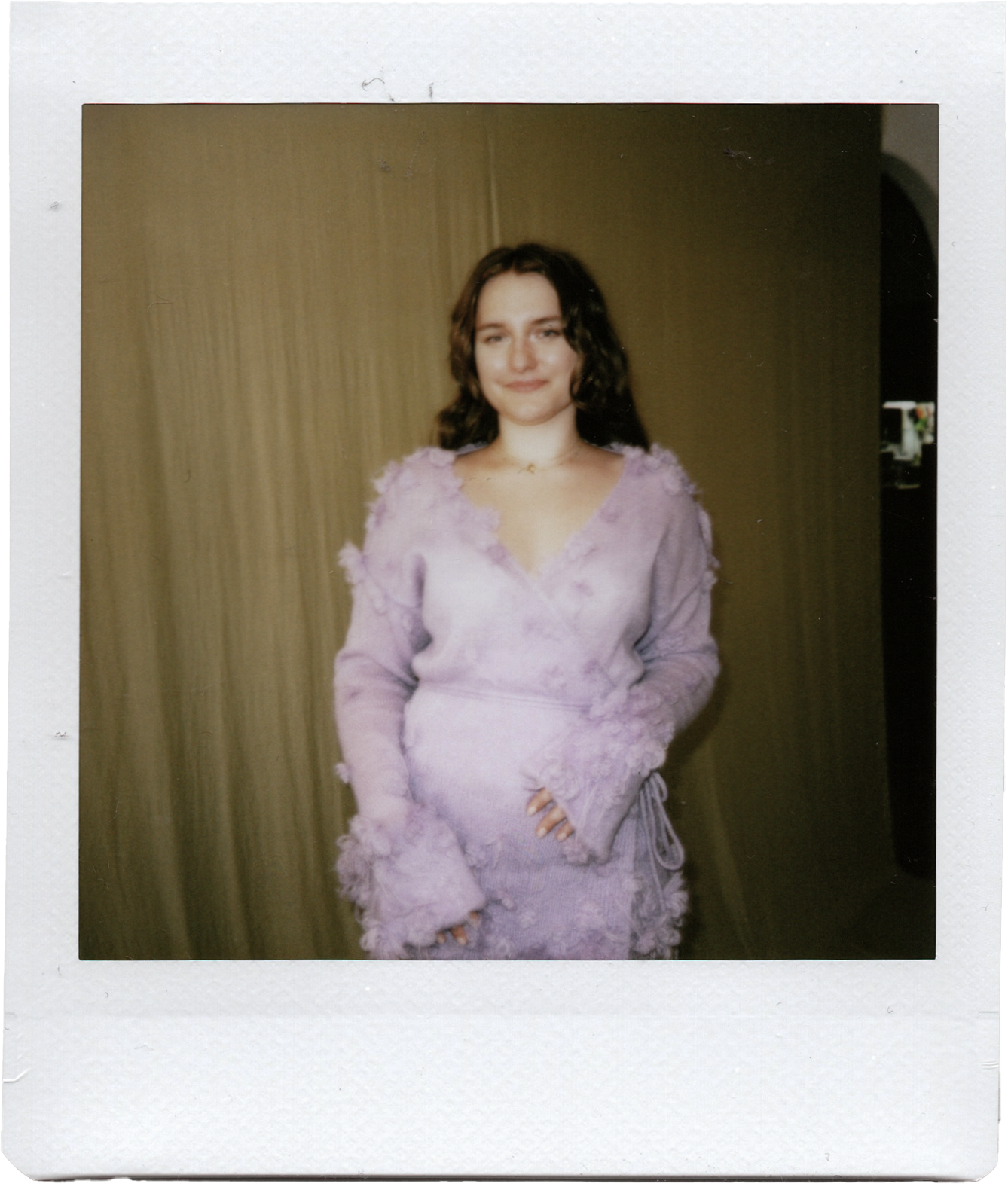 A woman with shoulder-length curly brown hair and a soft smile, wearing a lavender dress with textured and ruffled details, standing indoors against a neutral-colored wall.