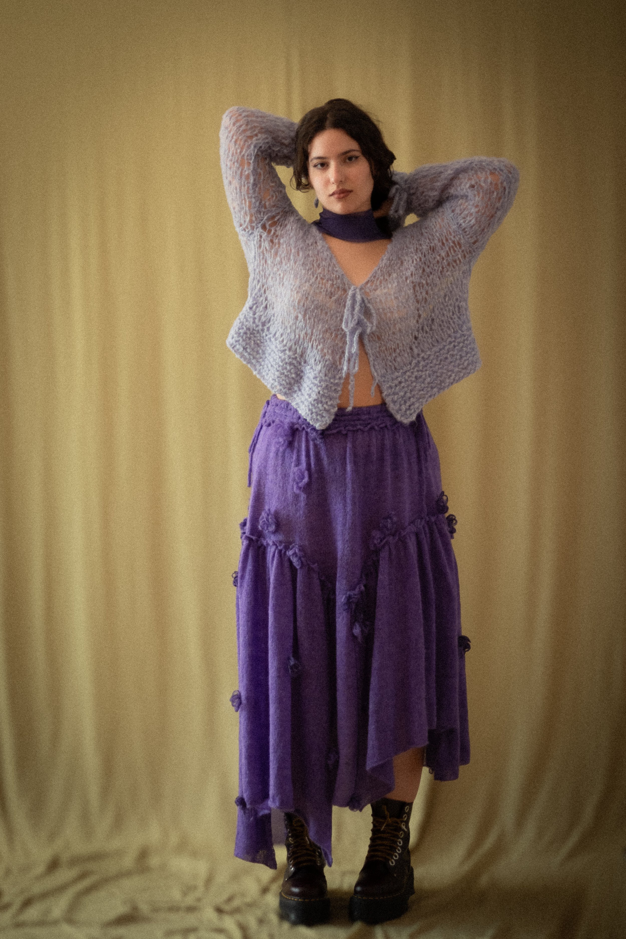 A woman with short dark hair wearing a purple dress, a light knit cardigan, and black boots, standing against a plain yellowish background with her hands behind her head.