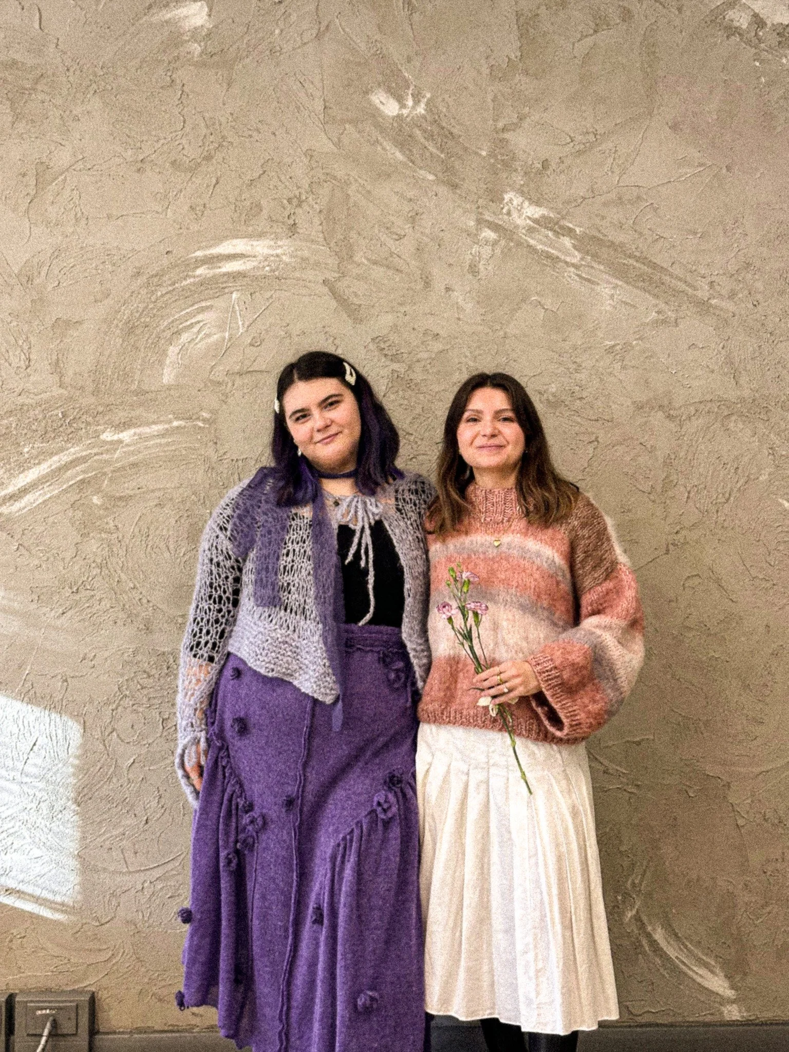Two women standing close together against a textured beige wall, one holding a small bouquet of pink flowers.