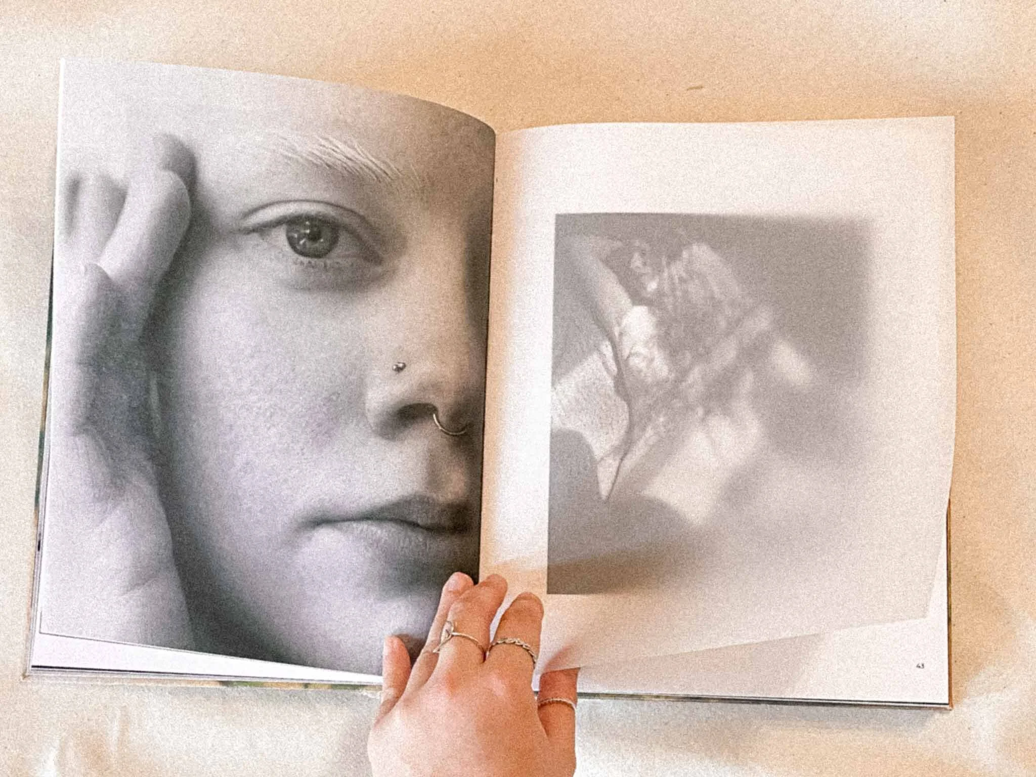 Open book featuring a black and white photograph of a woman's face with a piercing below her nose and a finger with rings holding the page.