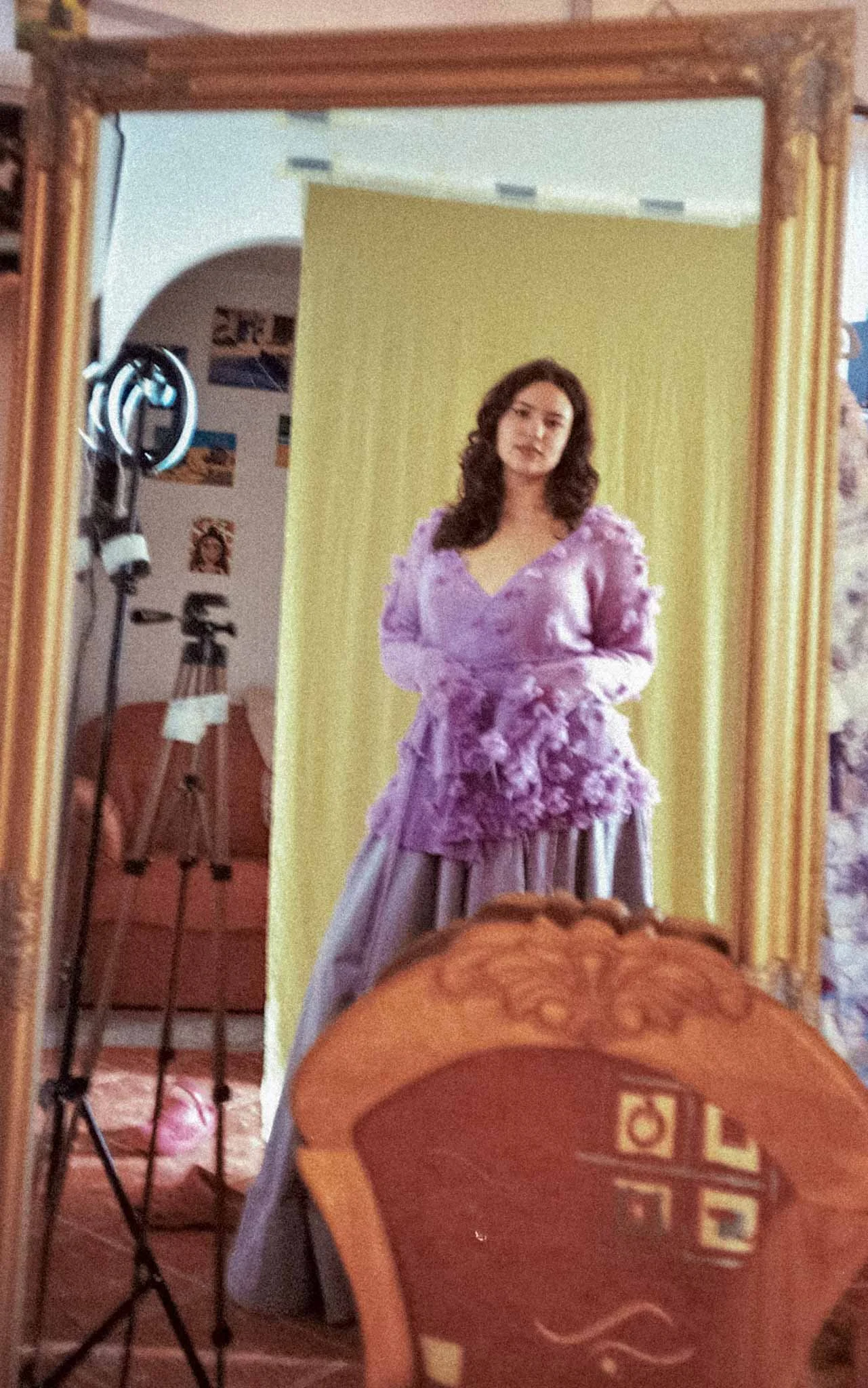 A woman with dark, wavy hair is taking a mirror selfie. She is wearing a purple dress with floral embellishments on the top and a long skirt. Behind her, there is a yellow backdrop, and various studio equipment can be seen in the background, including a chair, a tripod, and lighting equipment.