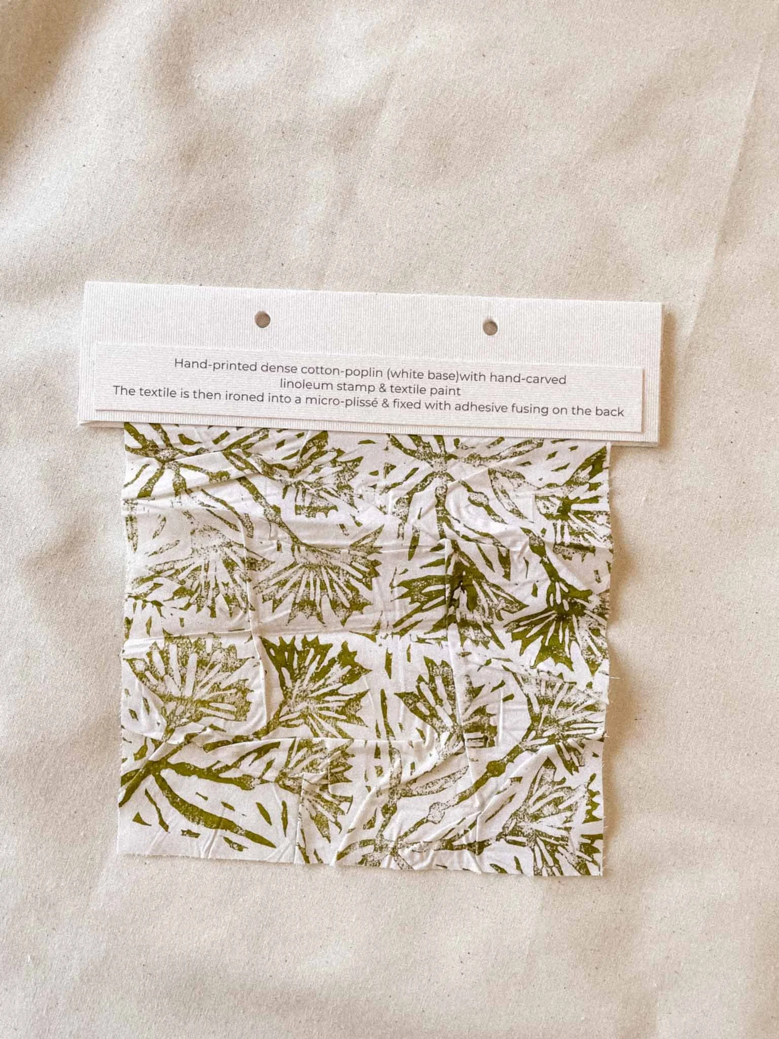 Close-up of fabric with a tag describing hand-printed dense cotton poplin with hand-carved linoleum stamp and textile paint, ironed into a micro-pliissé and fixed with adhesive fusings.
