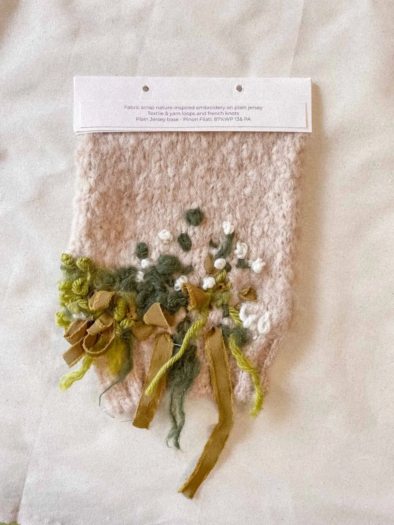 A fabric scrap with embroidery on plain jersey, featuring a mix of yarn loops, French knots, and ribbons in various colors including green, white, and brown.