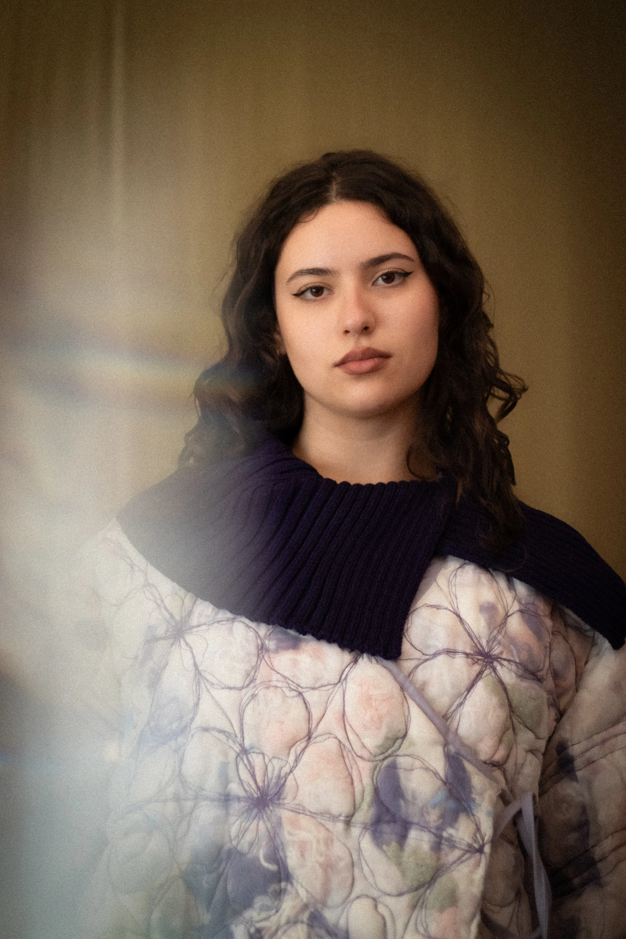 A young woman with wavy dark hair and winged eyeliner posing for a photo, wearing a dark sweater.