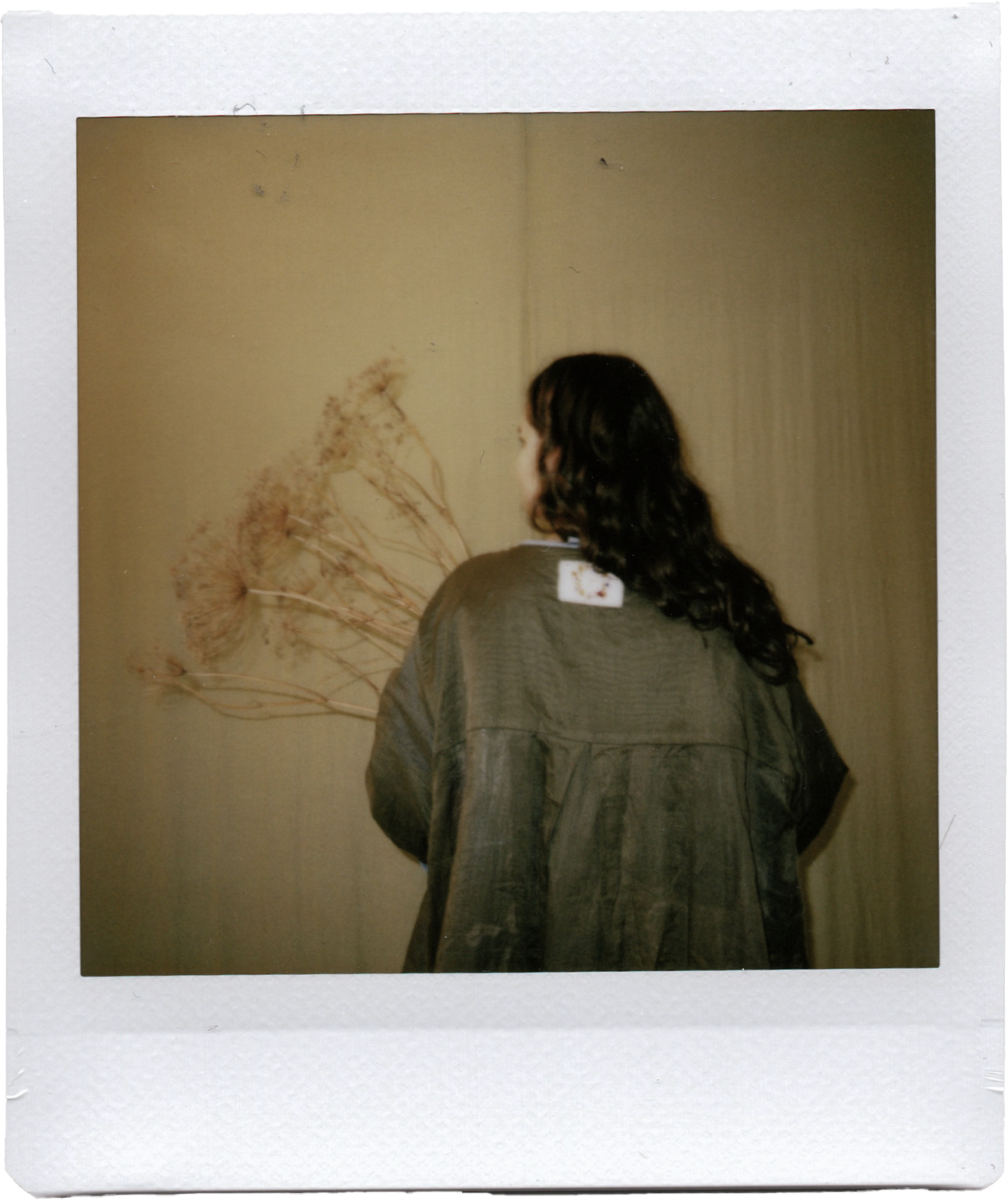 A woman with dark, wavy hair wearing a green jacket standing in front of a wall with dried flowers hanging on it