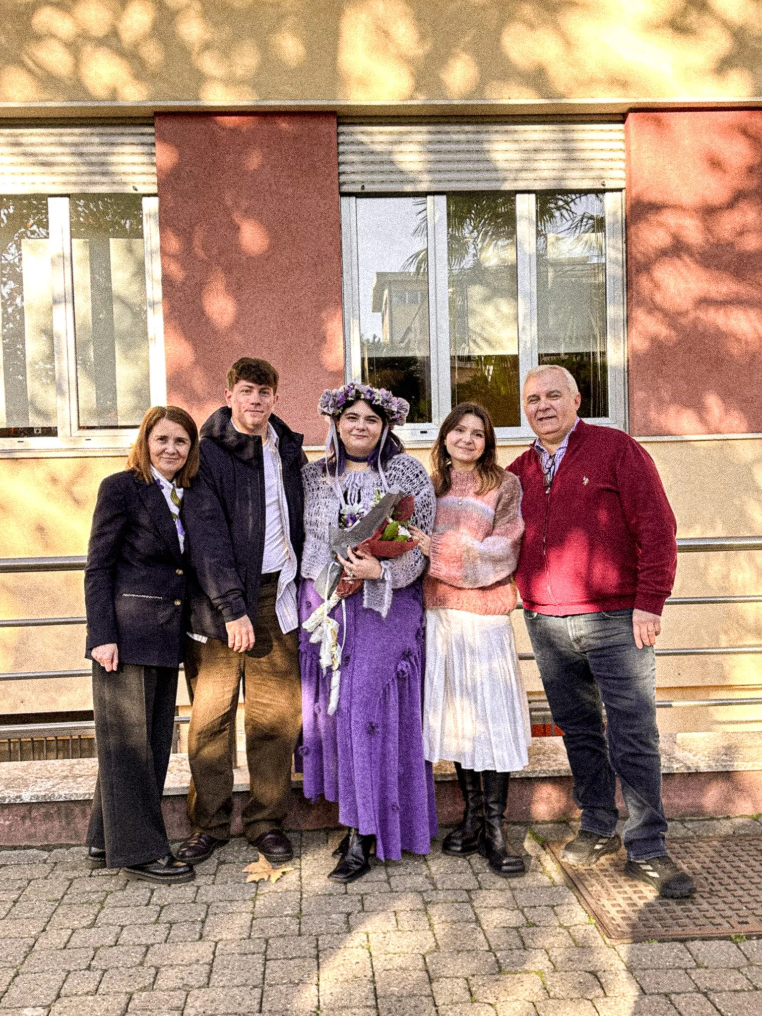 Group of five people standing outdoors in front of a building with large windows, during sunset or late afternoon, with trees and sunlight reflections visible in the windows. One woman in the center is holding a bouquet of flowers and wearing a purpl