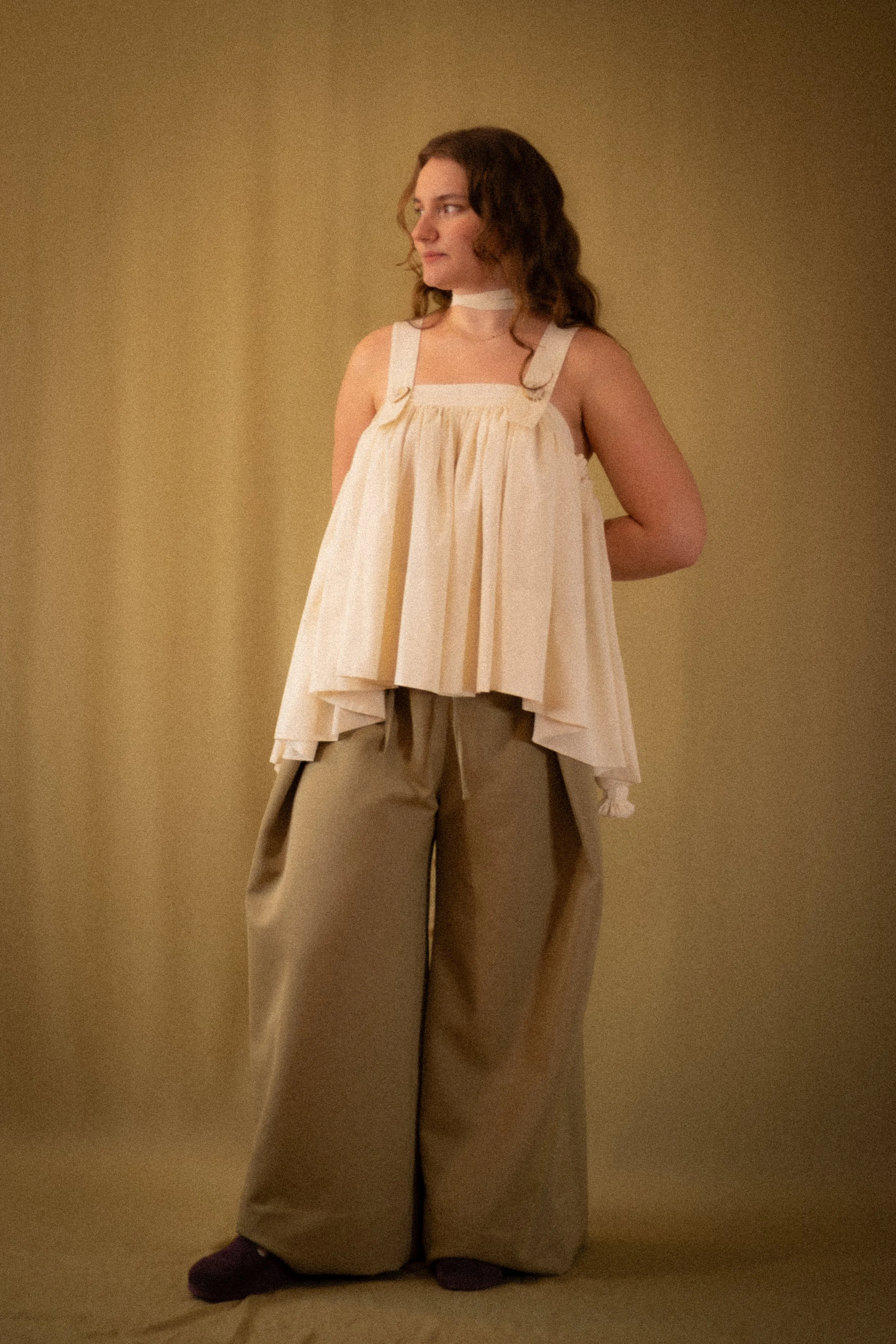 A woman with brown hair wearing a cream-colored sleeveless top with a square neckline and loose, wide-leg tan pants, standing against a plain beige background.