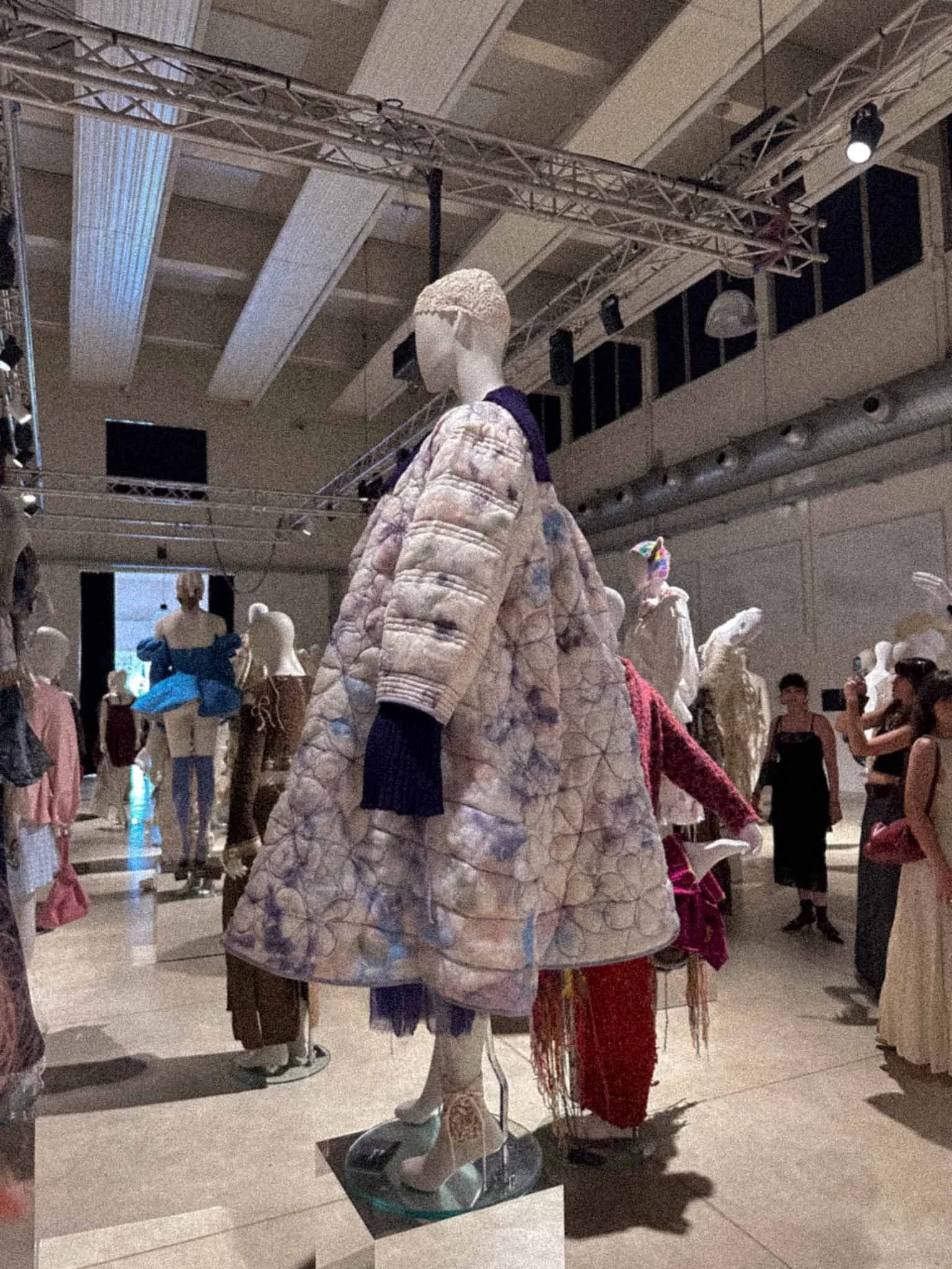 A mannequin dressed in a quilted, patterned coat with a large collar, displayed in a museum or gallery with other mannequins and visitors in the background.
