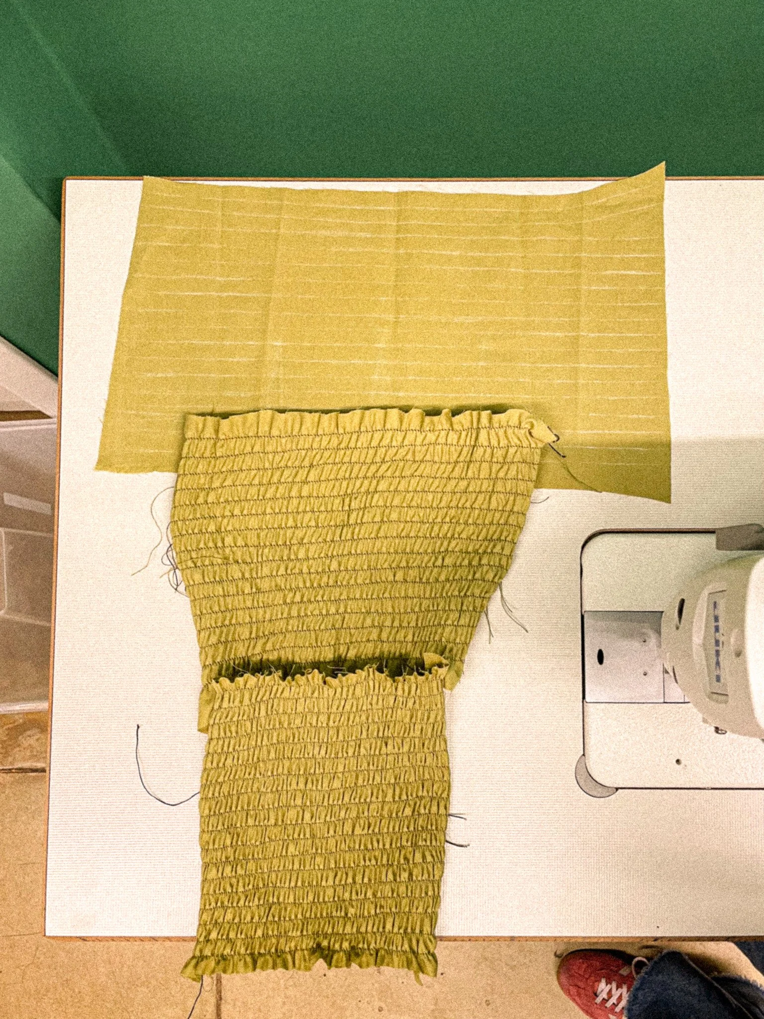Yellow fabric item on a white surface with a green wall background and a desk to the right, including some electronic devices and objects, with a person's foot wearing a red sneaker visible at the bottom right corner.