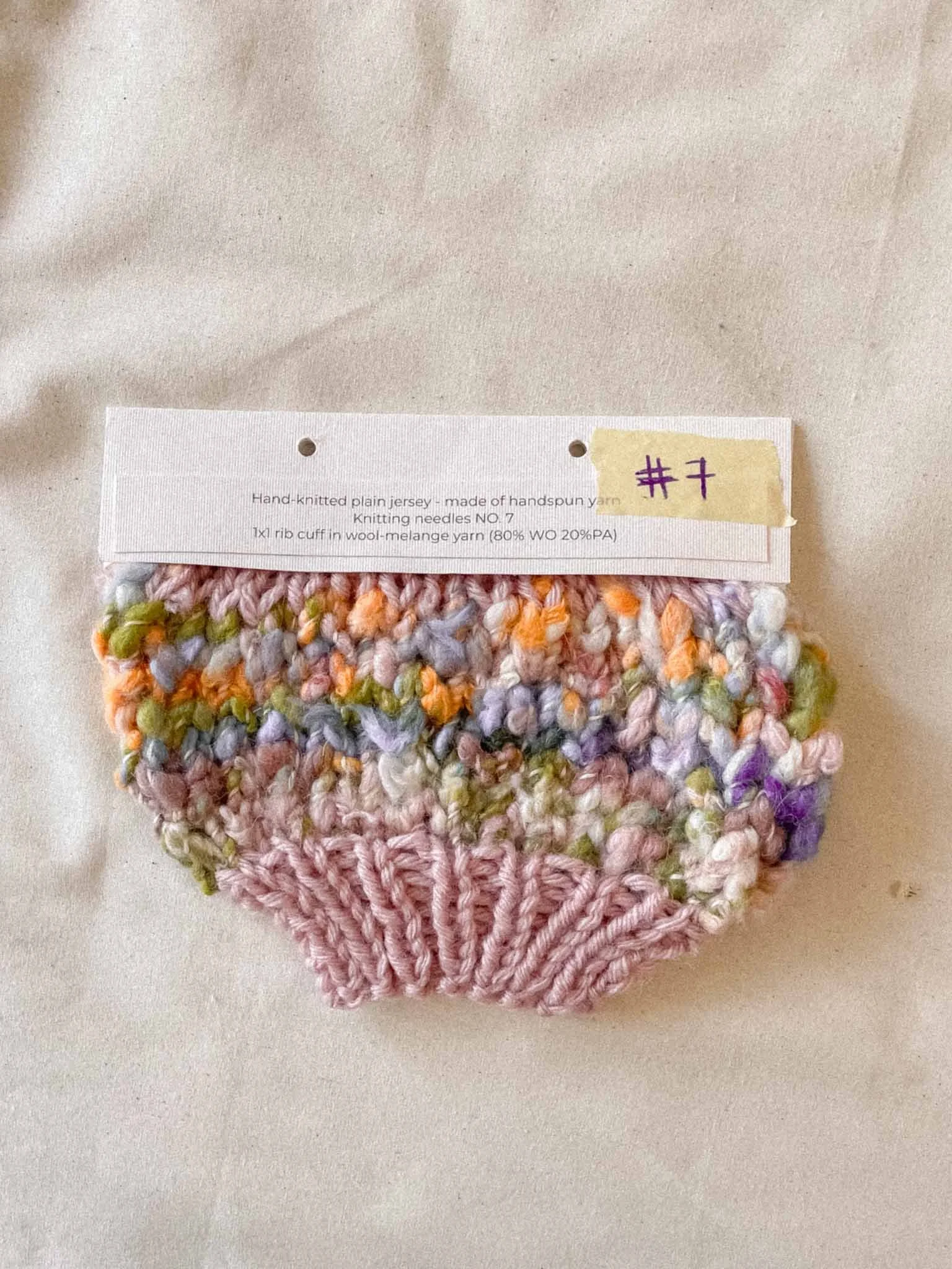 Colorful, hand-knitted yarn swatch with pastel shades of pink, purple, orange, and green, attached to a white tag that reads "#7" and has a description about yarn and knitting needles.
