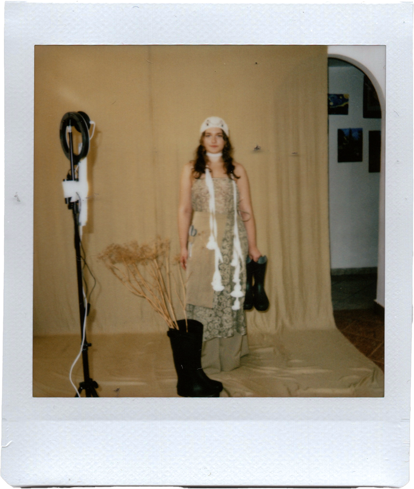 A woman standing indoors in front of a beige curtain background. She is wearing a white hat, a beige vest with various textures, tan pants, and black rain boots. She holds a pair of black rain boots in her right hand. There is a small black stand with dried plants on the left and an arched doorway to the right with a wall decorated with framed pictures.