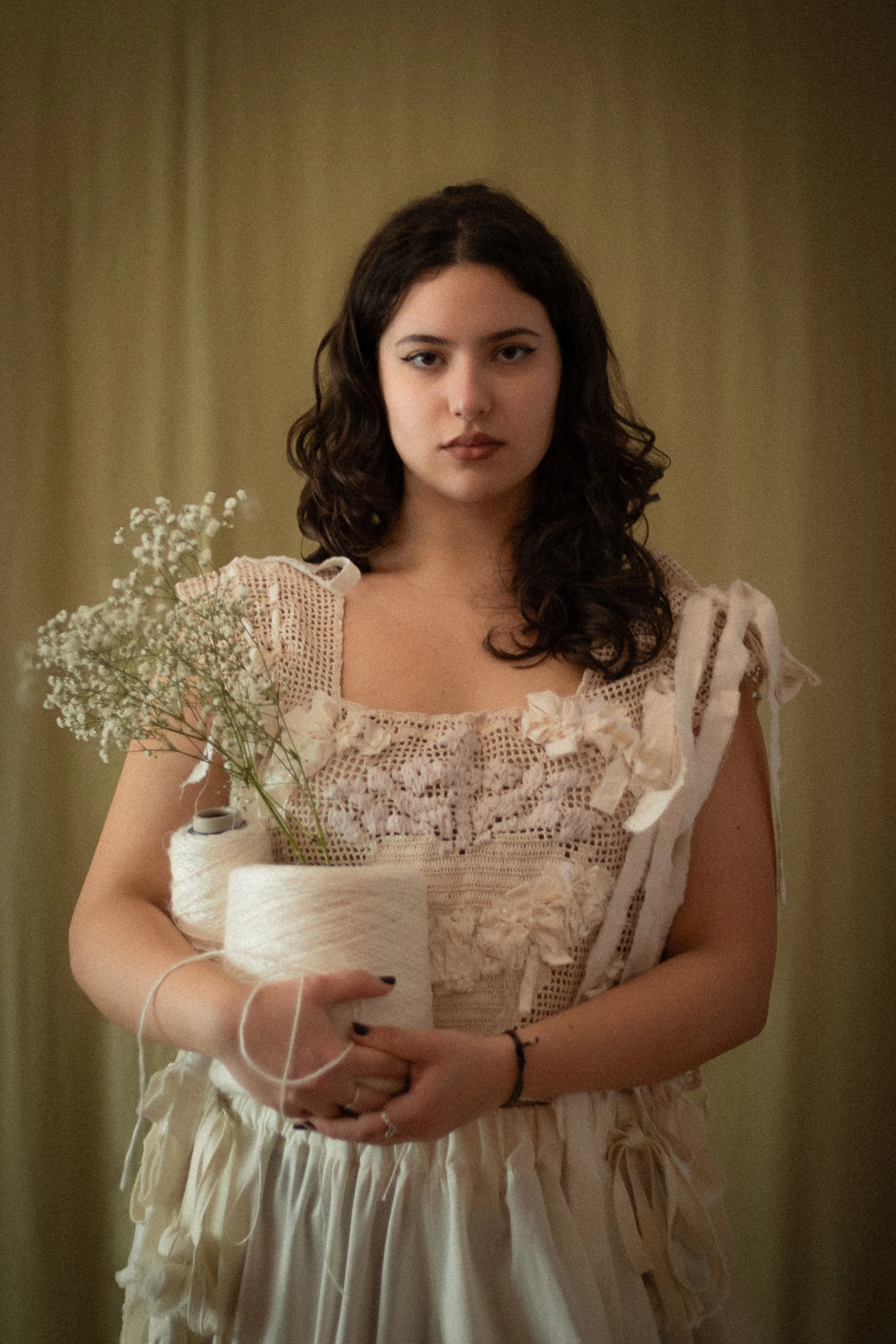 A woman with dark, curly hair and winged eyeliner holding a white bouquet with baby's breath flowers, wearing a vintage-style cream dress with lace and ruffled details, standing in front of a beige curtain.