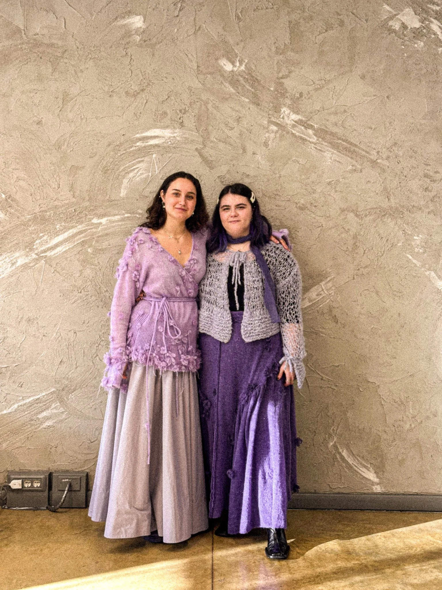 Two women pose together in front of a textured beige wall. They are dressed in elegant purple and lilac outfits, and both are smiling.