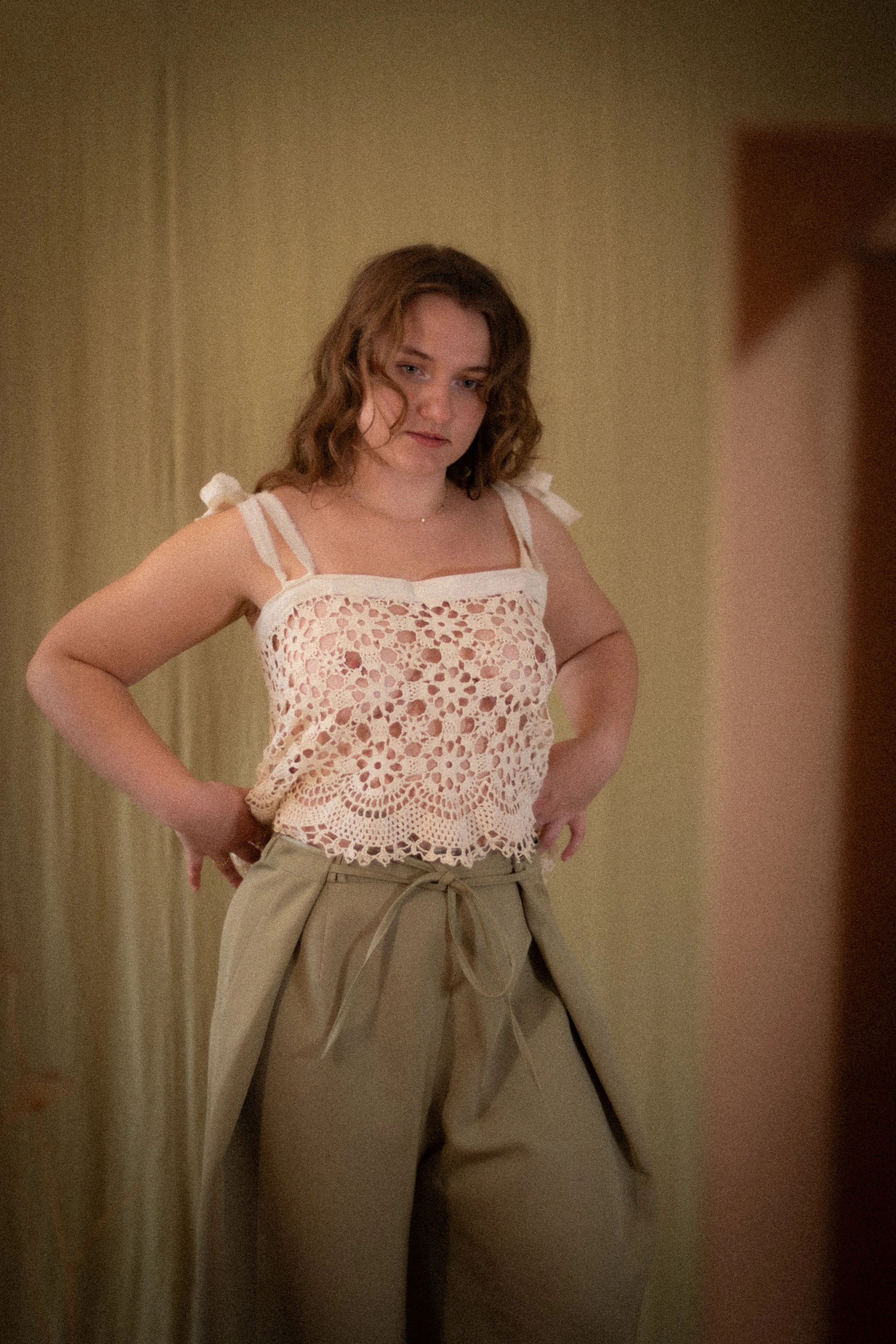 A woman with wavy, shoulder-length hair wearing a peach-colored, lacy tank top and beige high-waisted pants with a tie belt, standing in front of a beige curtain.