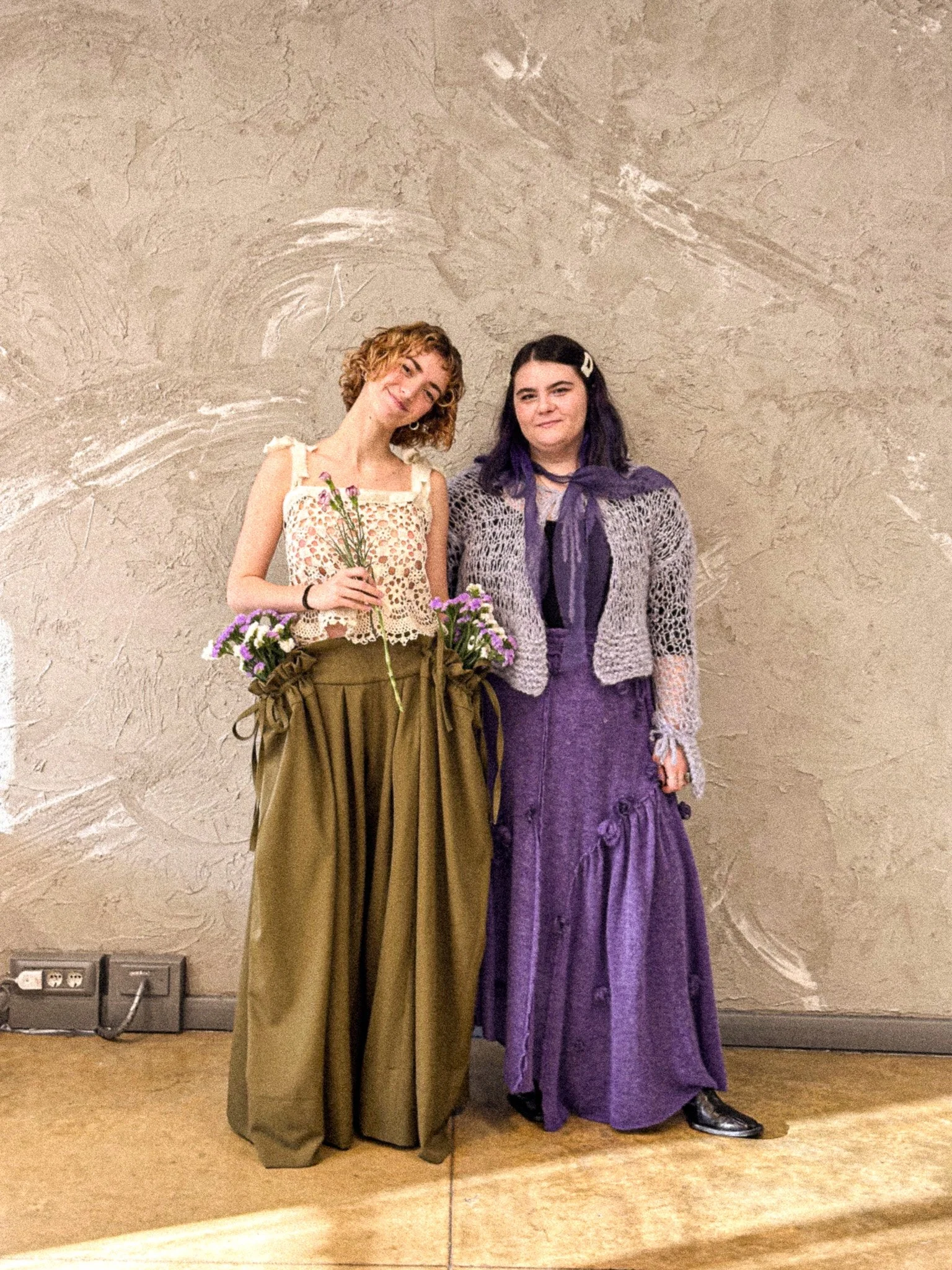 Two women standing together against a textured beige wall. The woman on the left has curly short blonde hair, holds a bouquet of flowers, and is wearing a lace top with beige and brown wide-leg pants. The woman on the right has dark hair with purple 