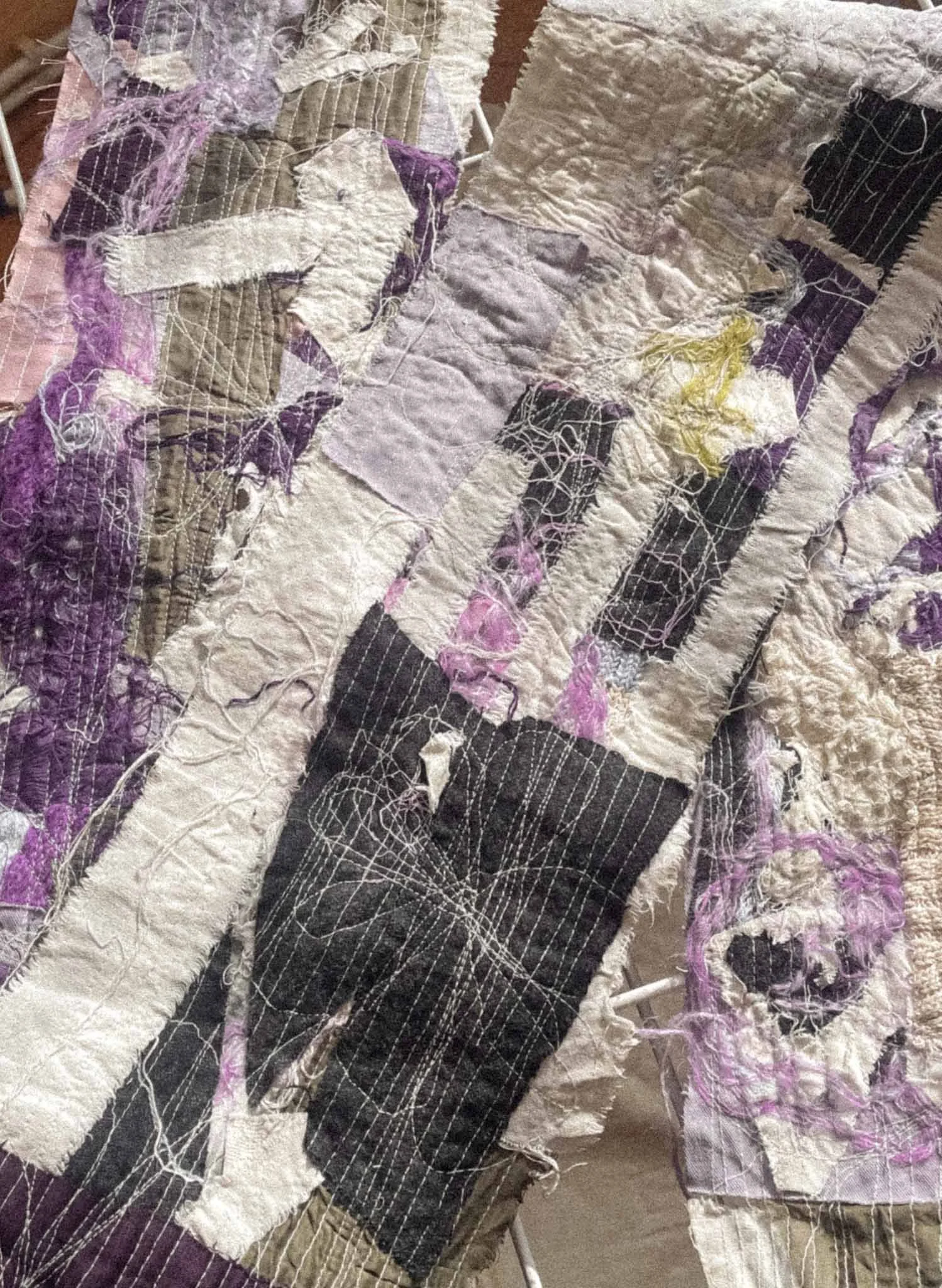 Close-up of a textured quilt with patches of fabric in various colors including purple, black, white, and beige, with visible embroidery, stitching, and frayed edges.