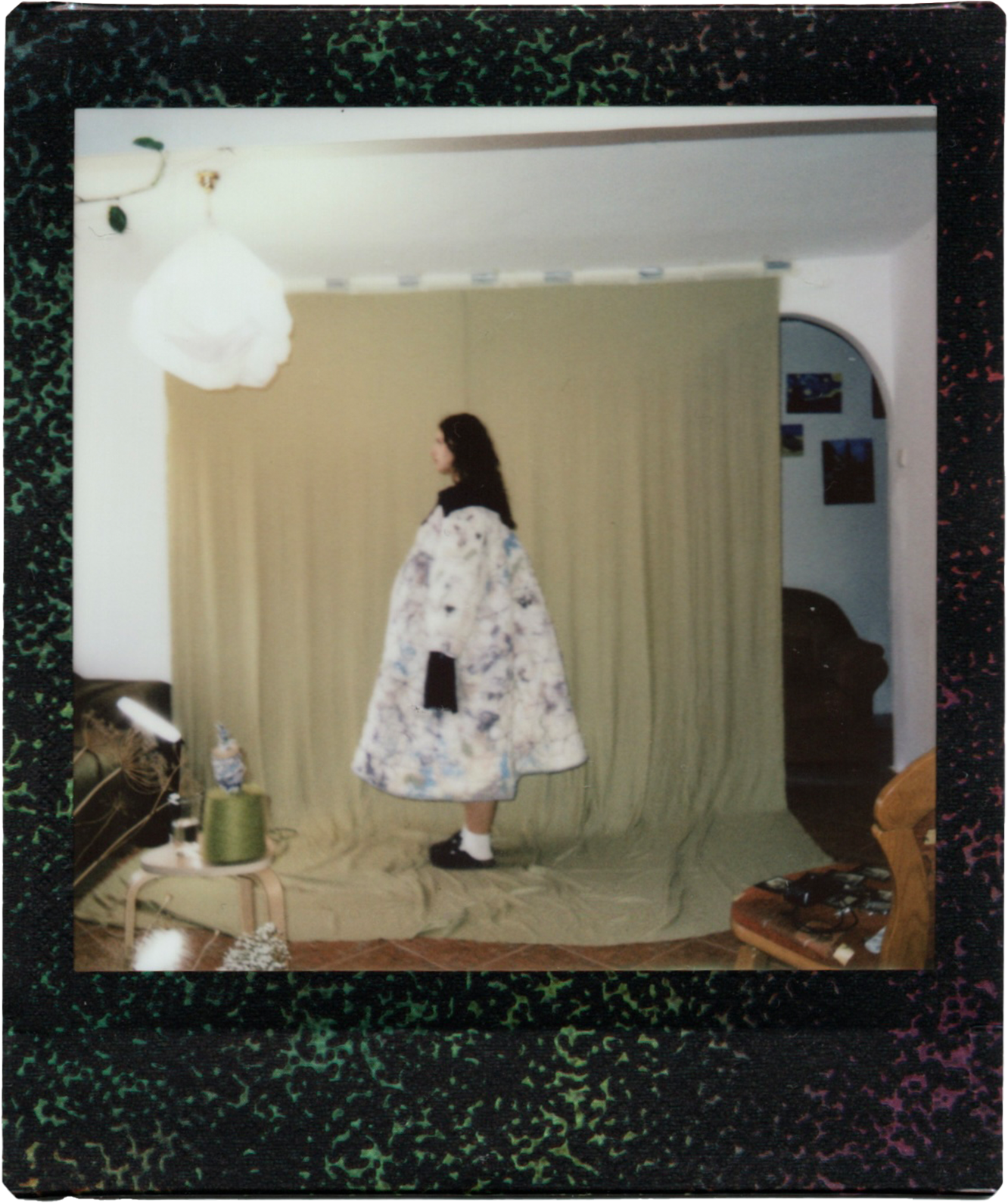 A woman standing sideways in a room with a yellow curtain background, wearing a floral coat, white socks, black shoes, and a black arm sleeve, surrounded by household items including a small table with a spool of green thread and a bottle, a chair, and photographs on the wall.