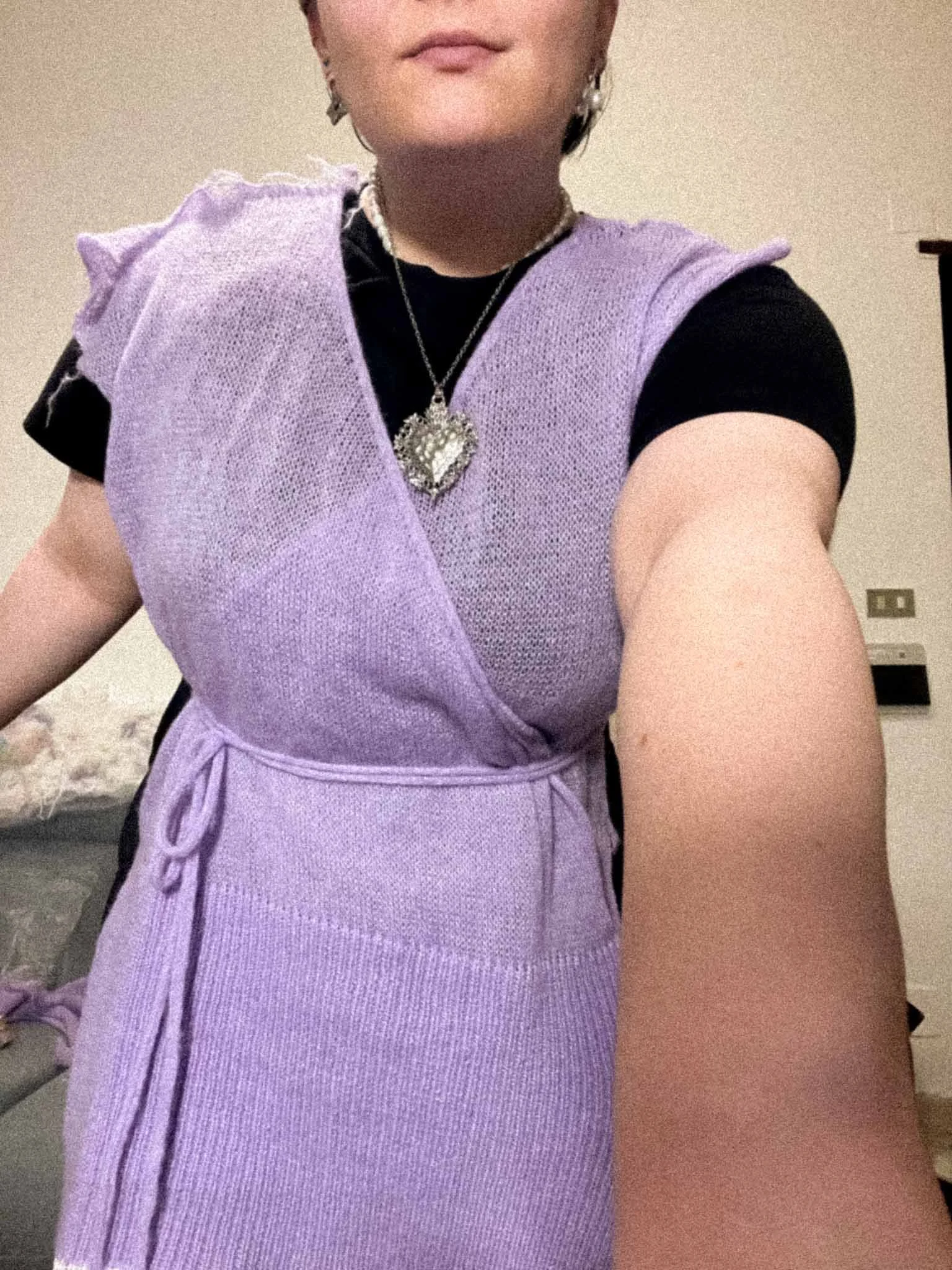 Person wearing a purple apron, black shirt, and jewelry, with only part of their face visible, smiling softly, indoors with a beige wall and electrical outlet in background.
