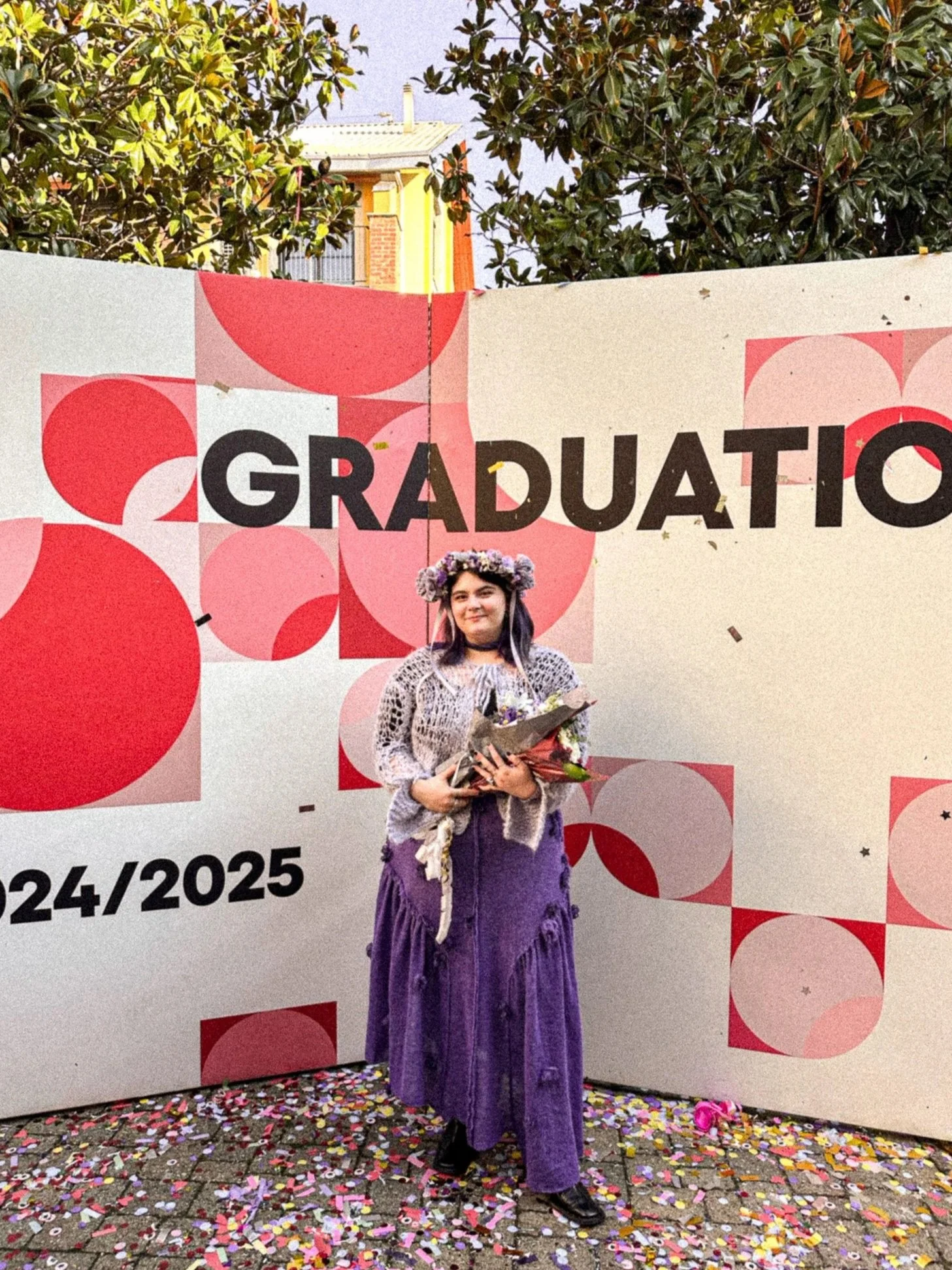A young woman dressed in purple with a flower crown, holding a bouquet and standing against a decorated backdrop that says 'GRADUATION' with the date '24/2025,' surrounded by colorful confetti on the ground.