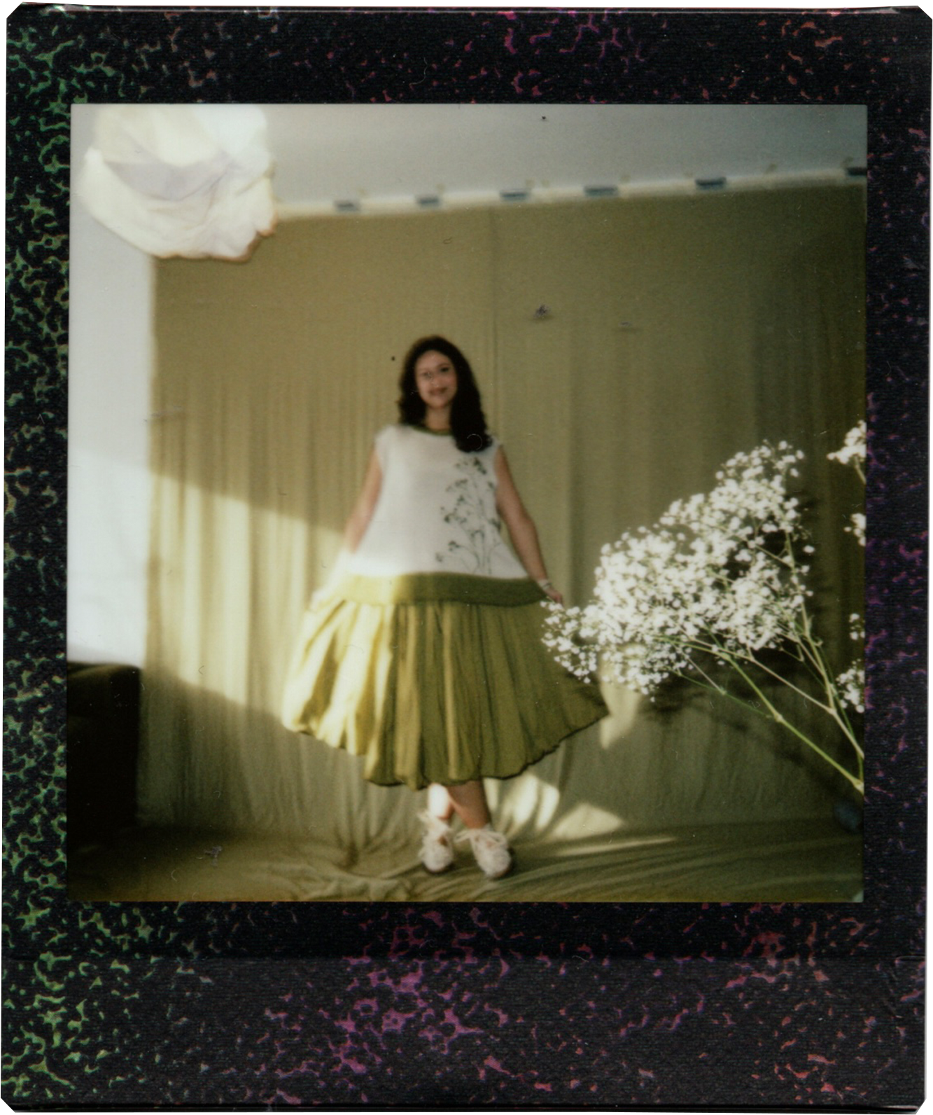 A young woman with dark hair and a light-colored top, standing indoors next to a large bouquet of white flowers, wearing a pleated yellow skirt, with a beige curtain behind her and a decorative border around the photo.