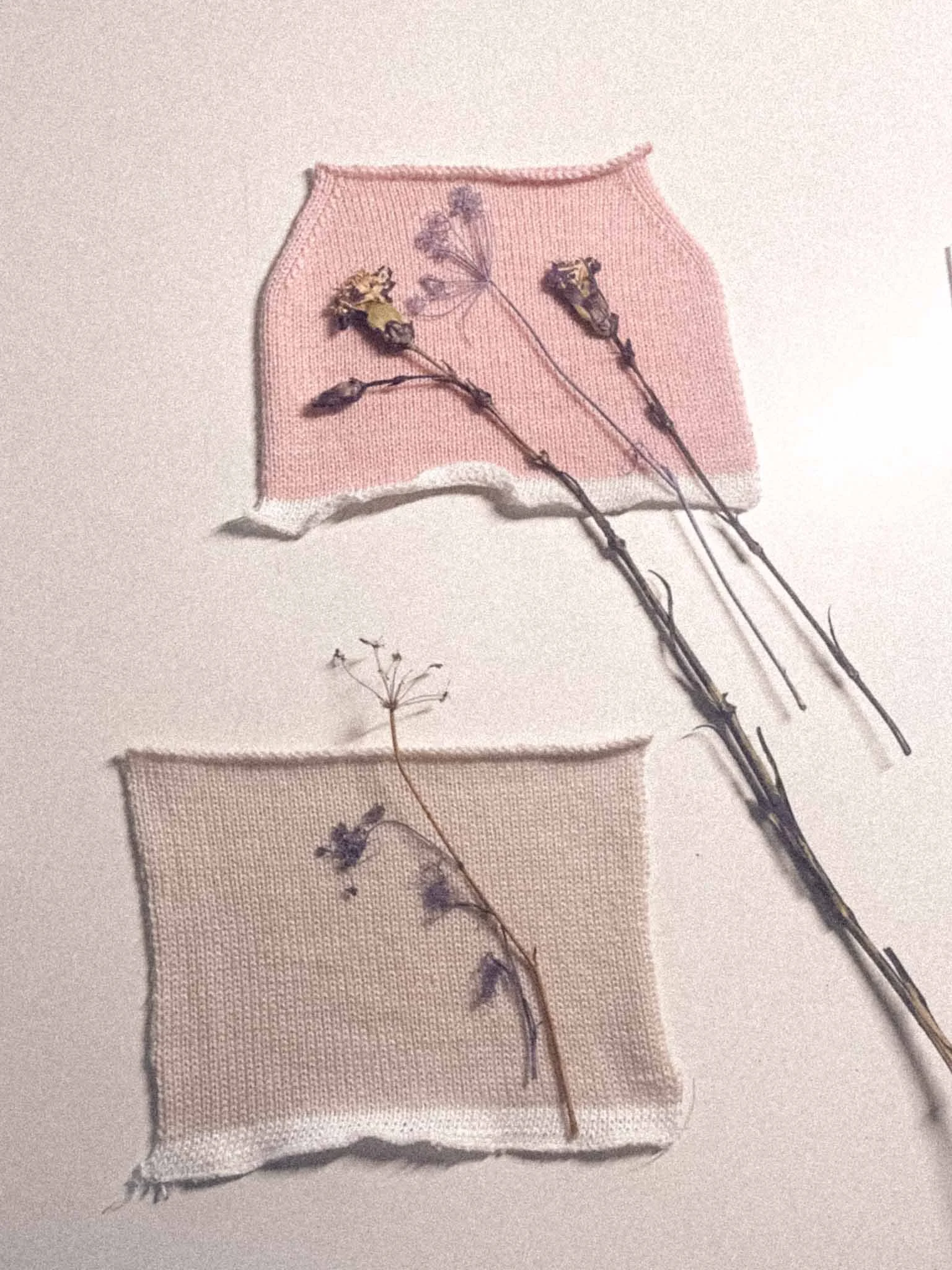 A pink knitted fabric square with a dried purple flower stitched onto it, and similar beige fabric square with a dried purple flower stitched onto it, arranged on a white surface.