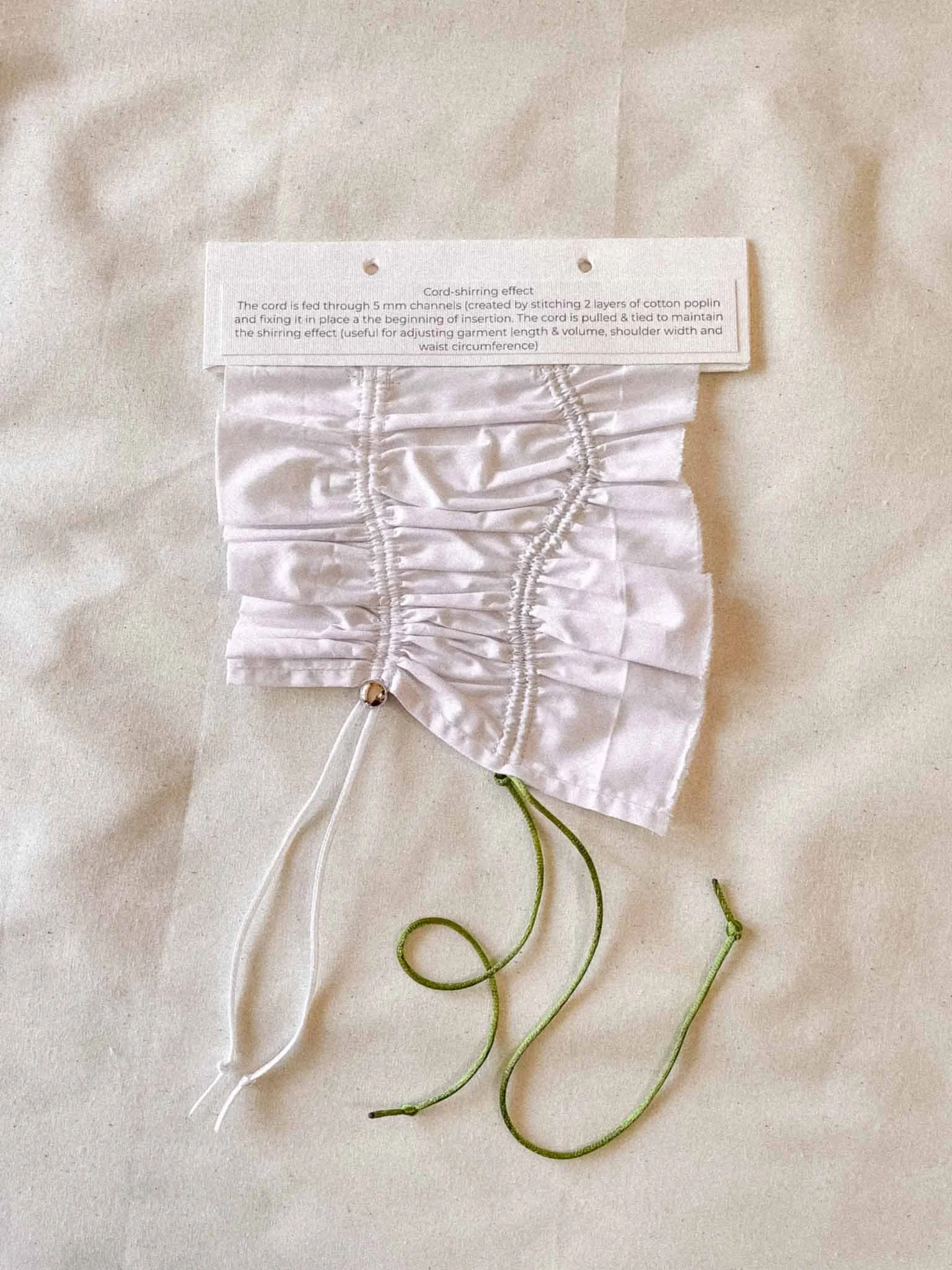 Close-up of a fabric face mask with a label attached, showing instructions for cord-shirring effect and green and white elastic cords on a light-colored background.