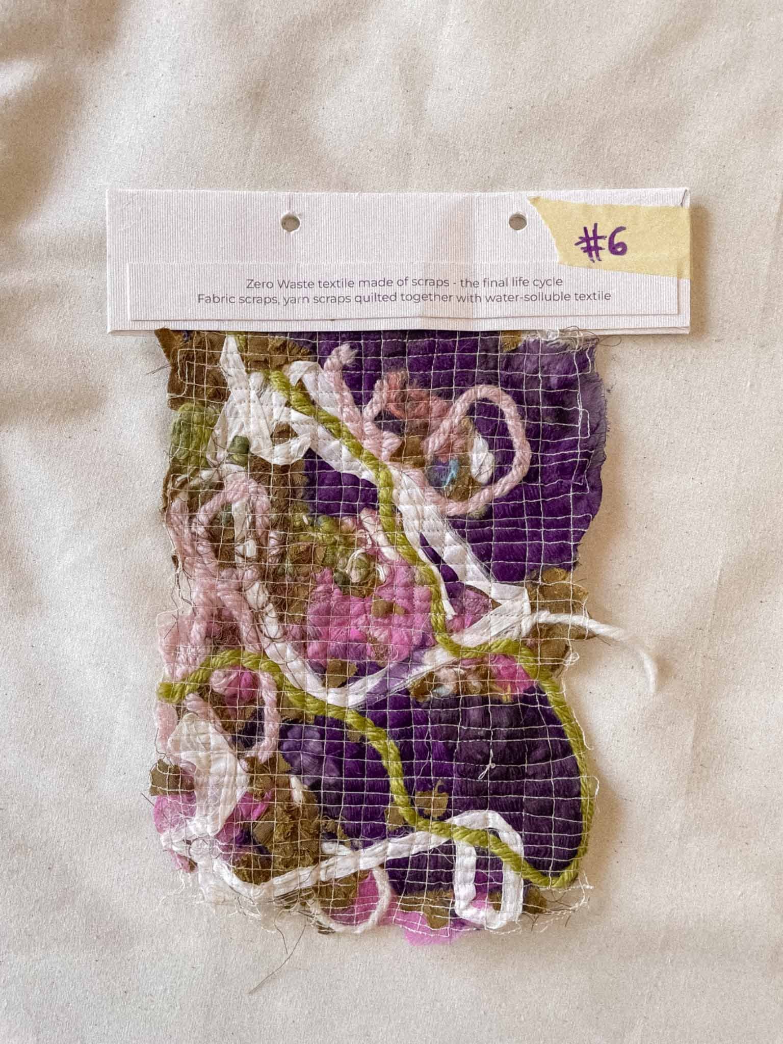 A piece of textile art made of assorted fabric scraps and yarn, wrapped in a white net, with a white label on top that reads 'Zero Waste textile made of scraps.' A yellow sticky note with '#6' is attached to the top right corner of the label.