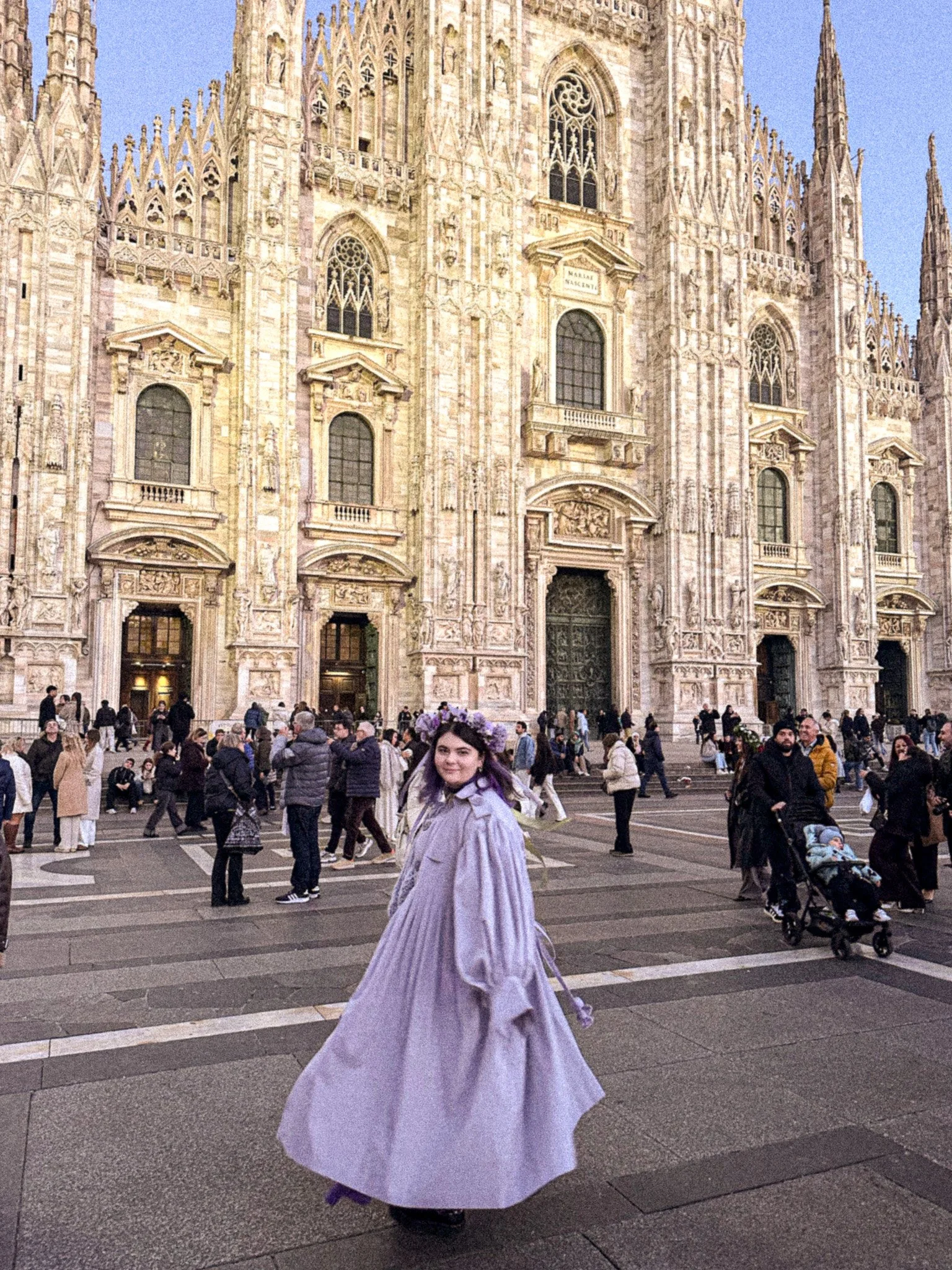 A woman in a flowing, lavender dress with a floral headpiece stands in a crowded plaza in front of a historic cathedral with intricate stone carvings and tall spires.