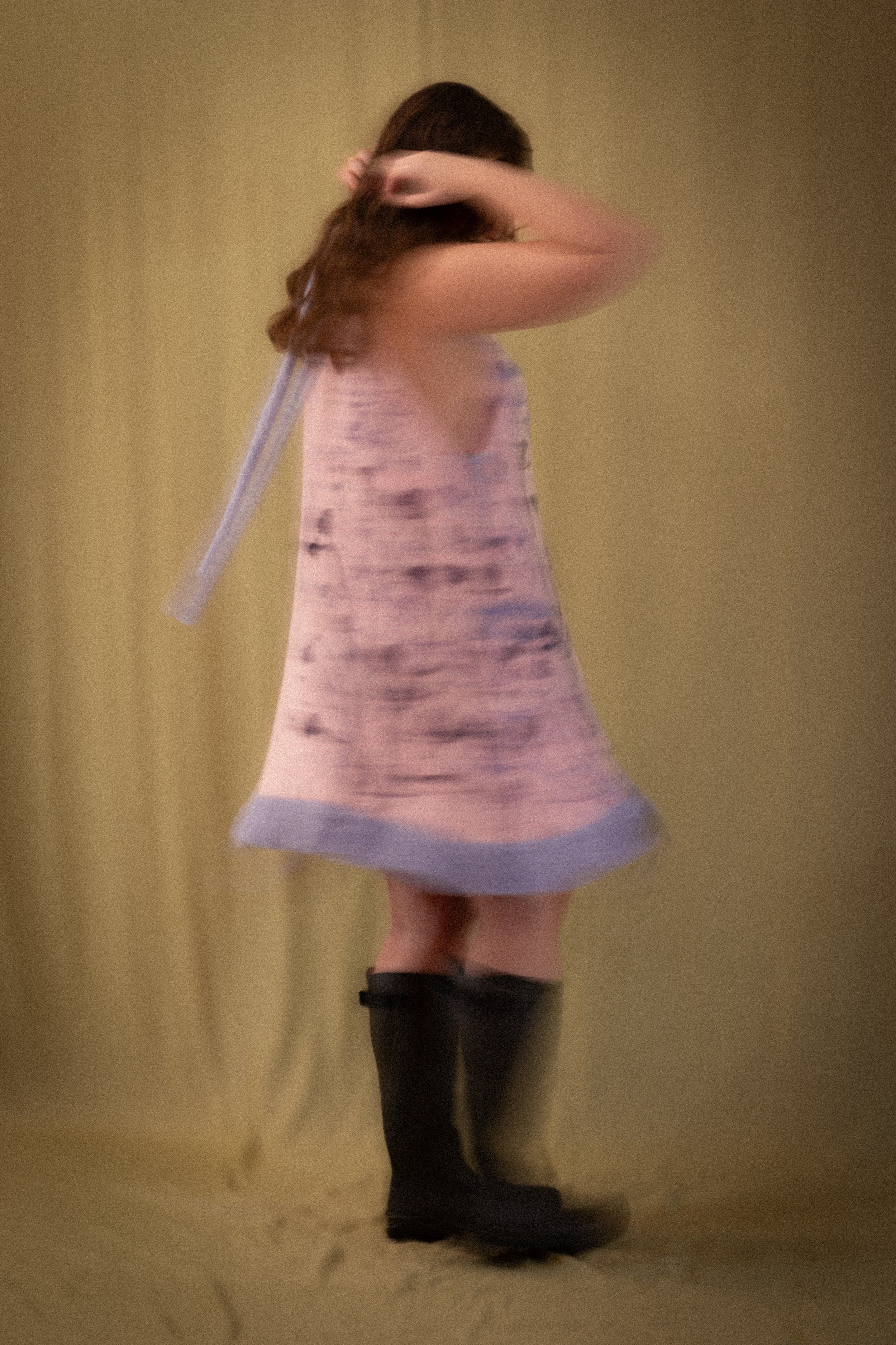 A woman wearing a sleeveless dress with a pastel-colored pattern and black thigh-high boots stands against a plain beige background, with one arm raised behind her head.