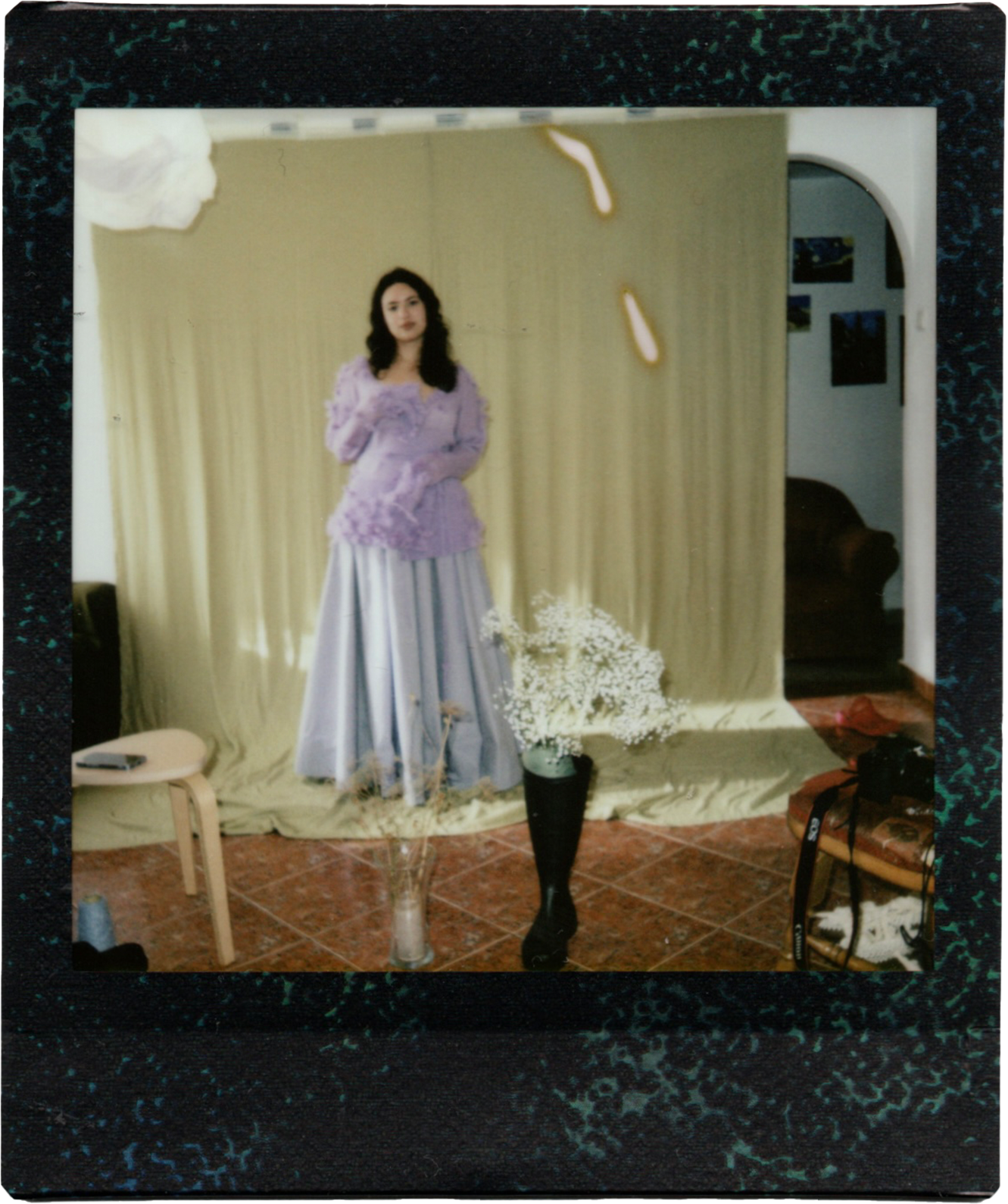 A woman dressed in a lavender top and a long, shiny silver skirt standing in front of a yellow curtain backdrop, with a bouquet of white baby's breath flowers in a tall vase in front of her.