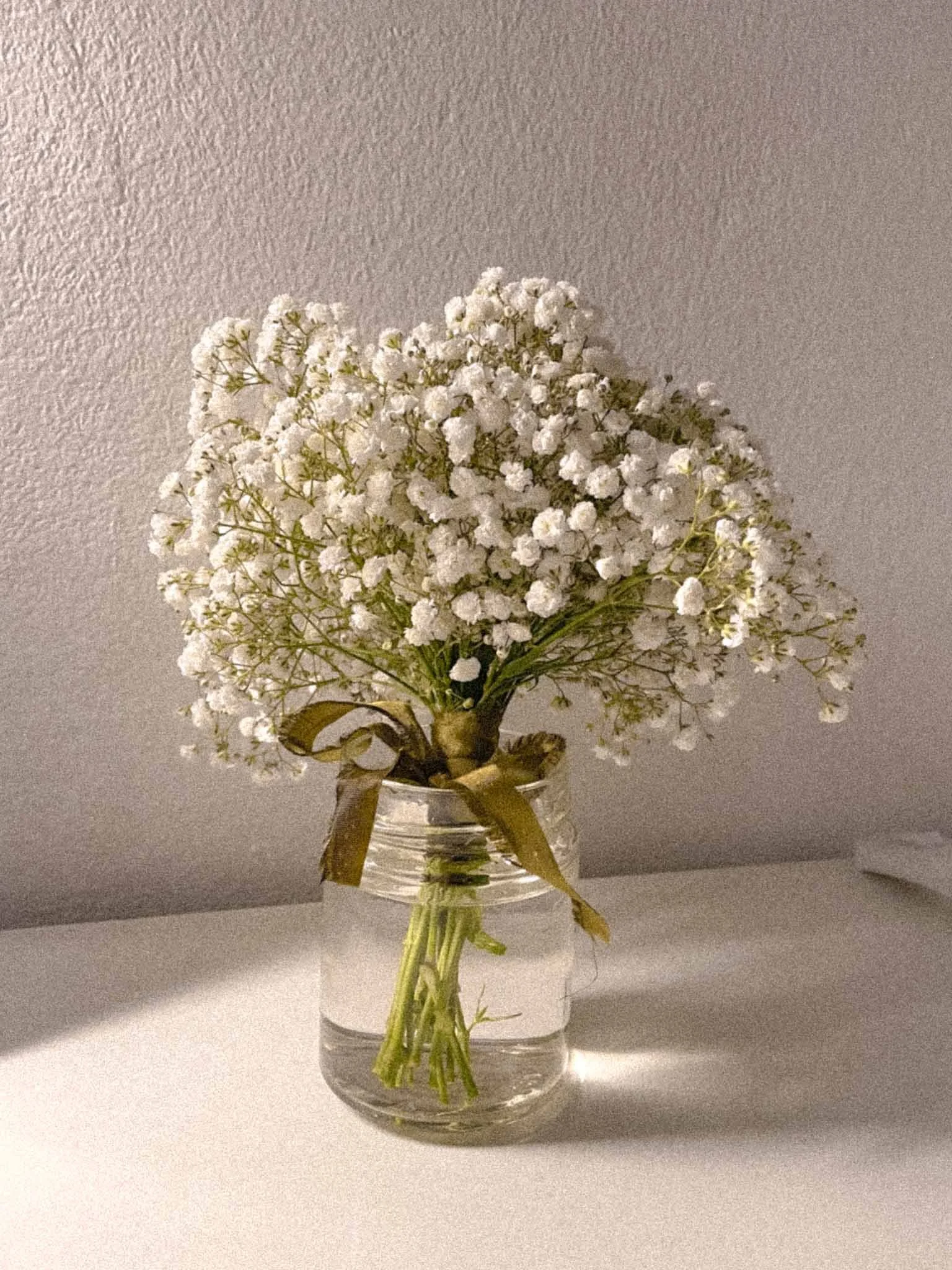 A bouquet of white baby’s breath flowers in a clear glass vase with a gold ribbon, placed on a white surface against a gray wall.