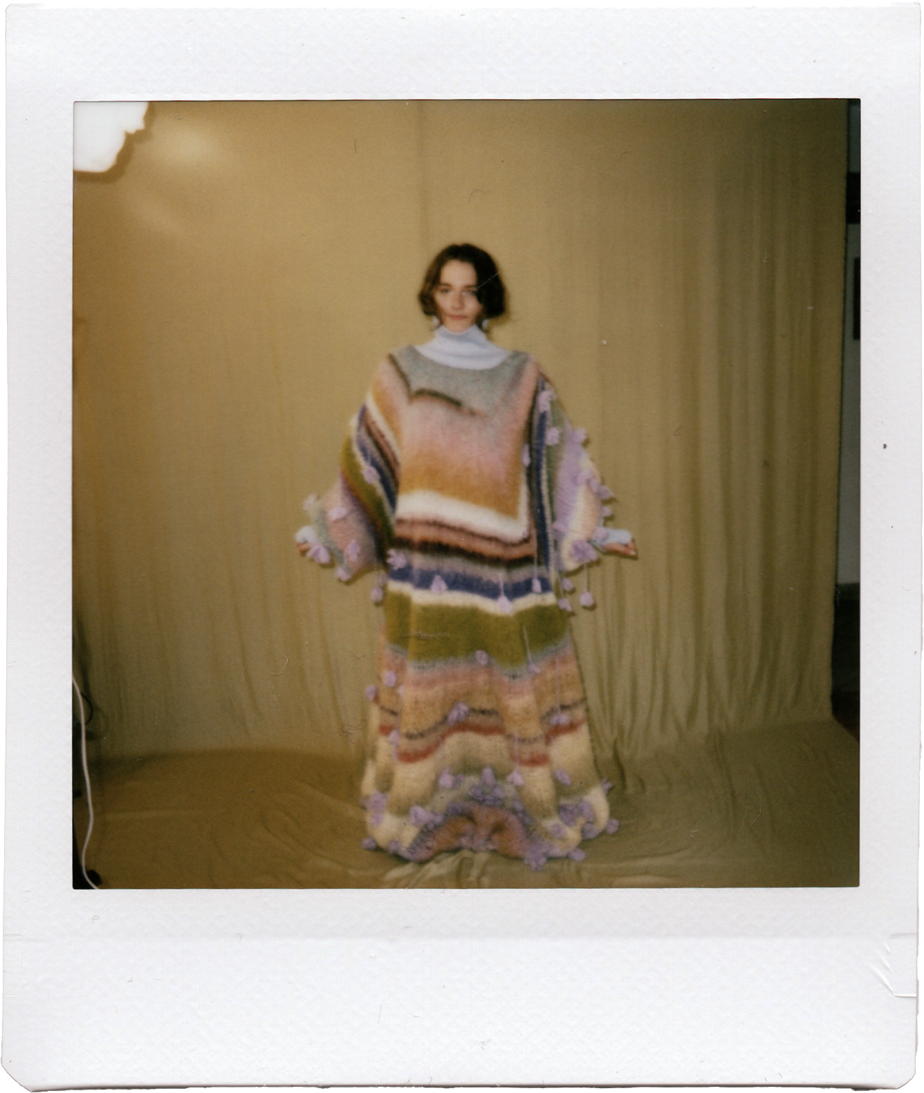 A young woman standing in front of a beige curtain, wearing a colorful, oversized knitted sweater and matching skirt, decorated with small purple pom-poms.