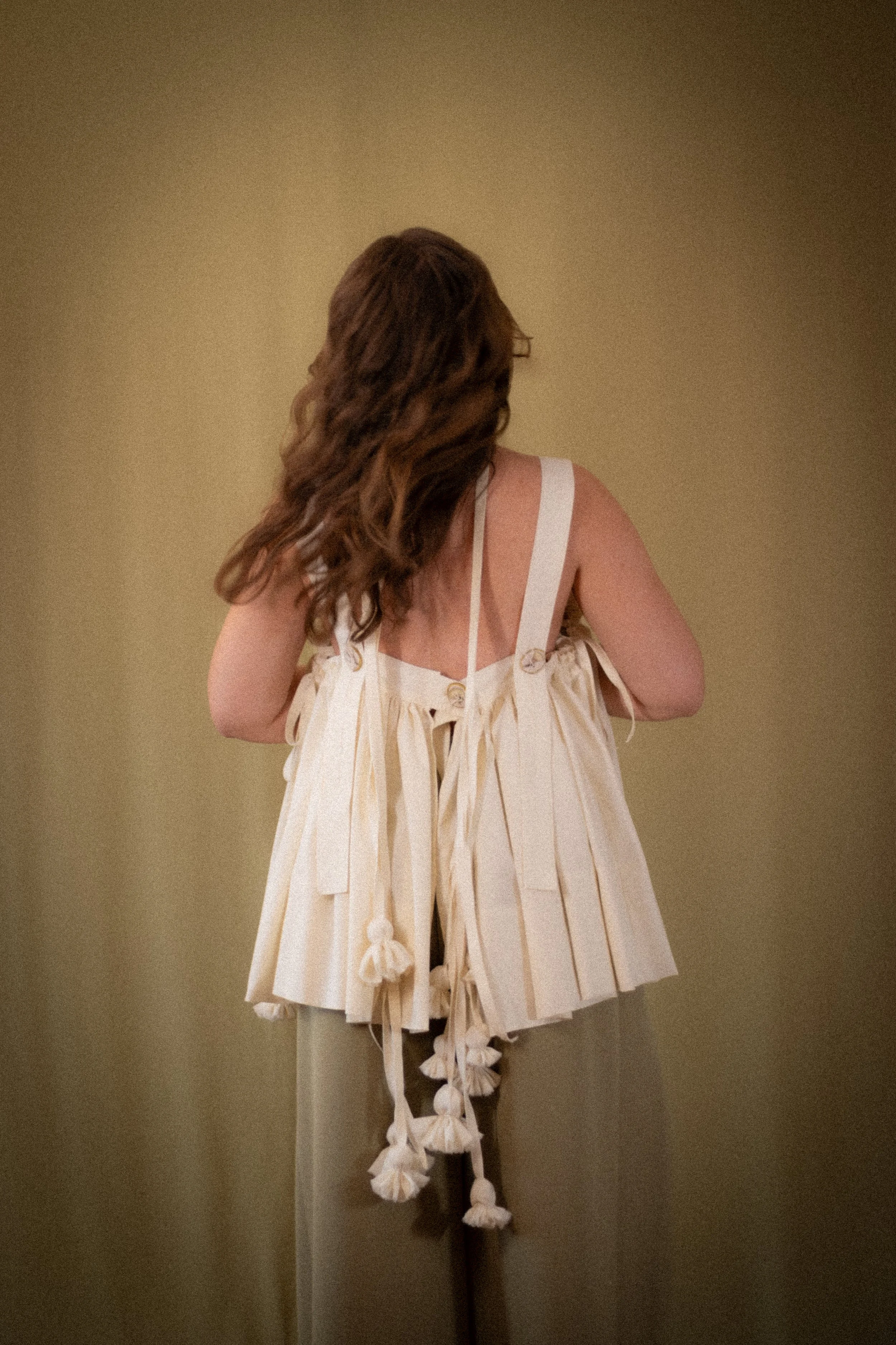 Back view of a woman with wavy brown hair wearing a white dress with ribbon straps and decorative tassels, standing against a beige wall.
