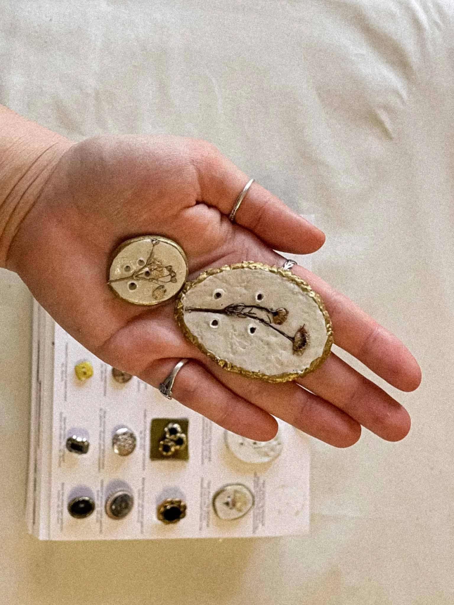 A person's hand with rings is holding two round, decorative buttons with dried plant inclusions. In the background, a display board shows a collection of various jewelry items, including rings and earrings, arranged in a grid.