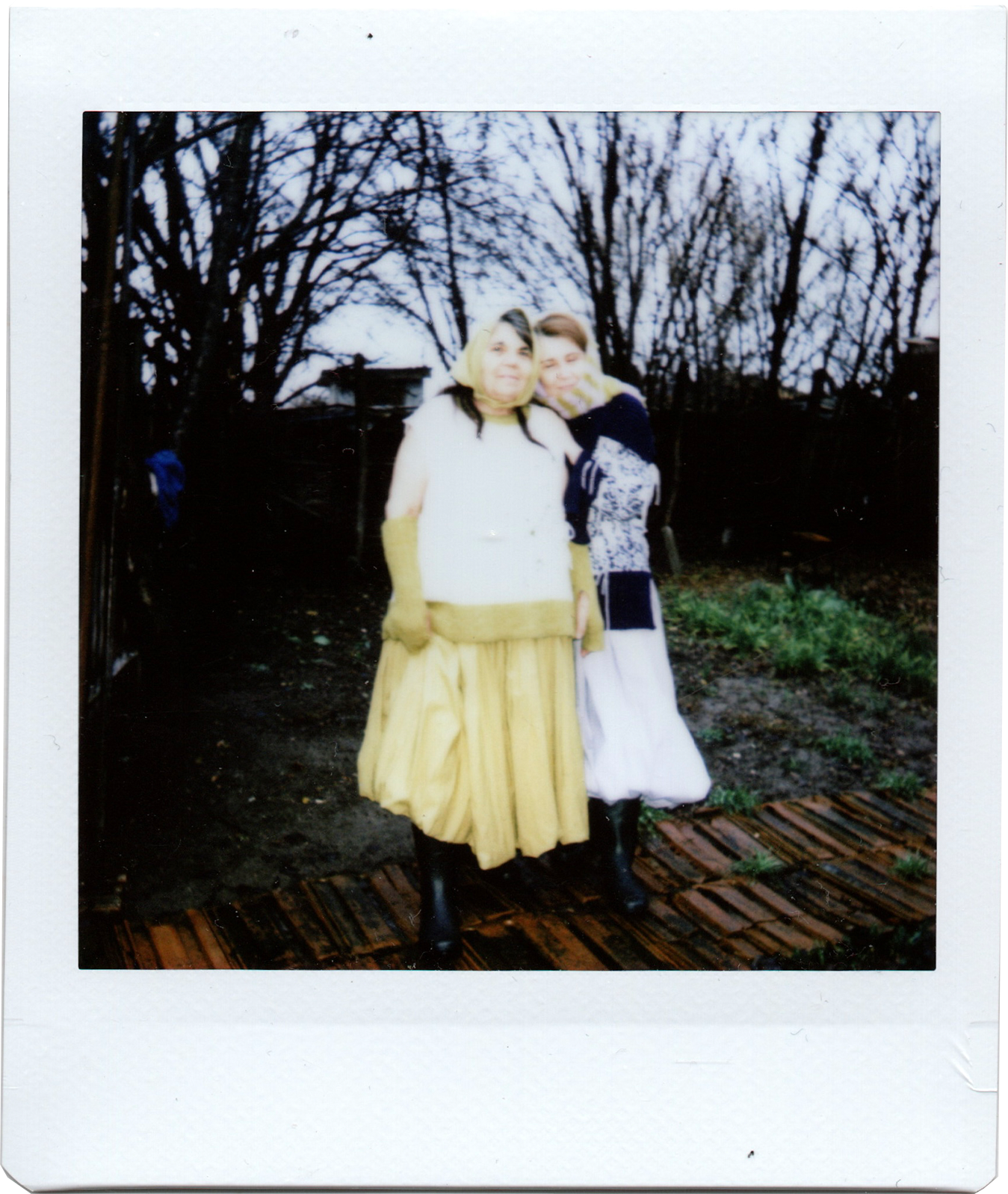 Two women standing outdoors on a wooden deck, dressed in vintage-style clothing with long skirts and sweaters, with leafless trees in the background on a cloudy day.