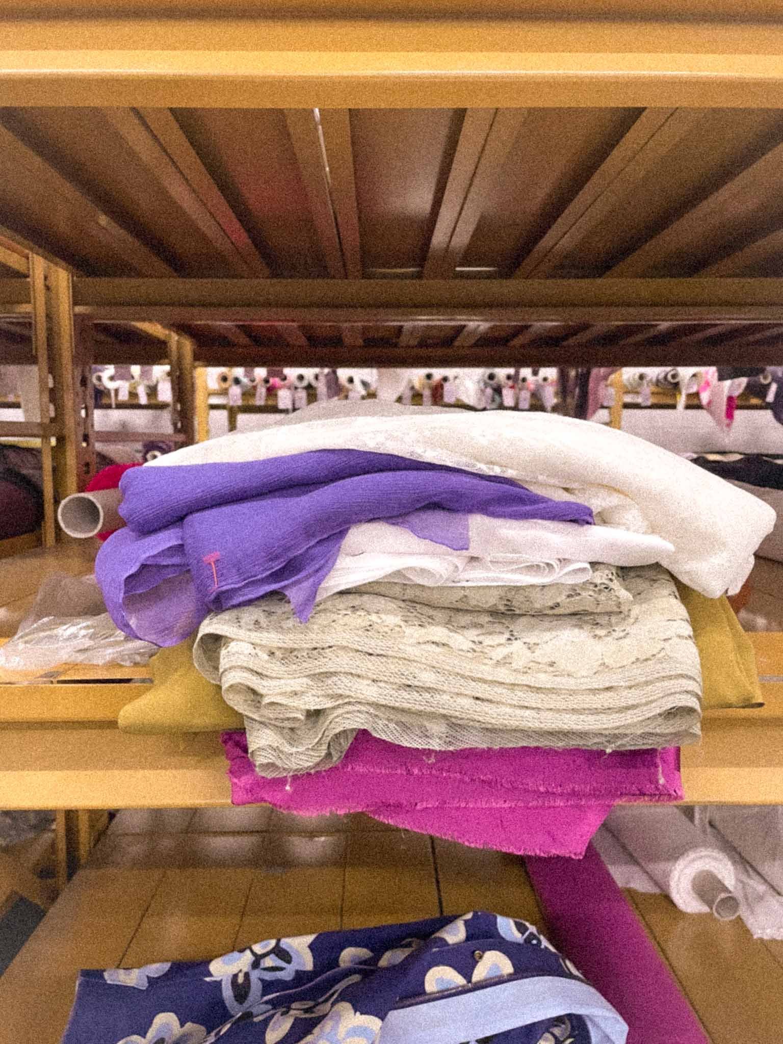 Stacked folded clothes and fabric on a table beneath a wooden shelf with rolls of paper and laundry room items in the background.