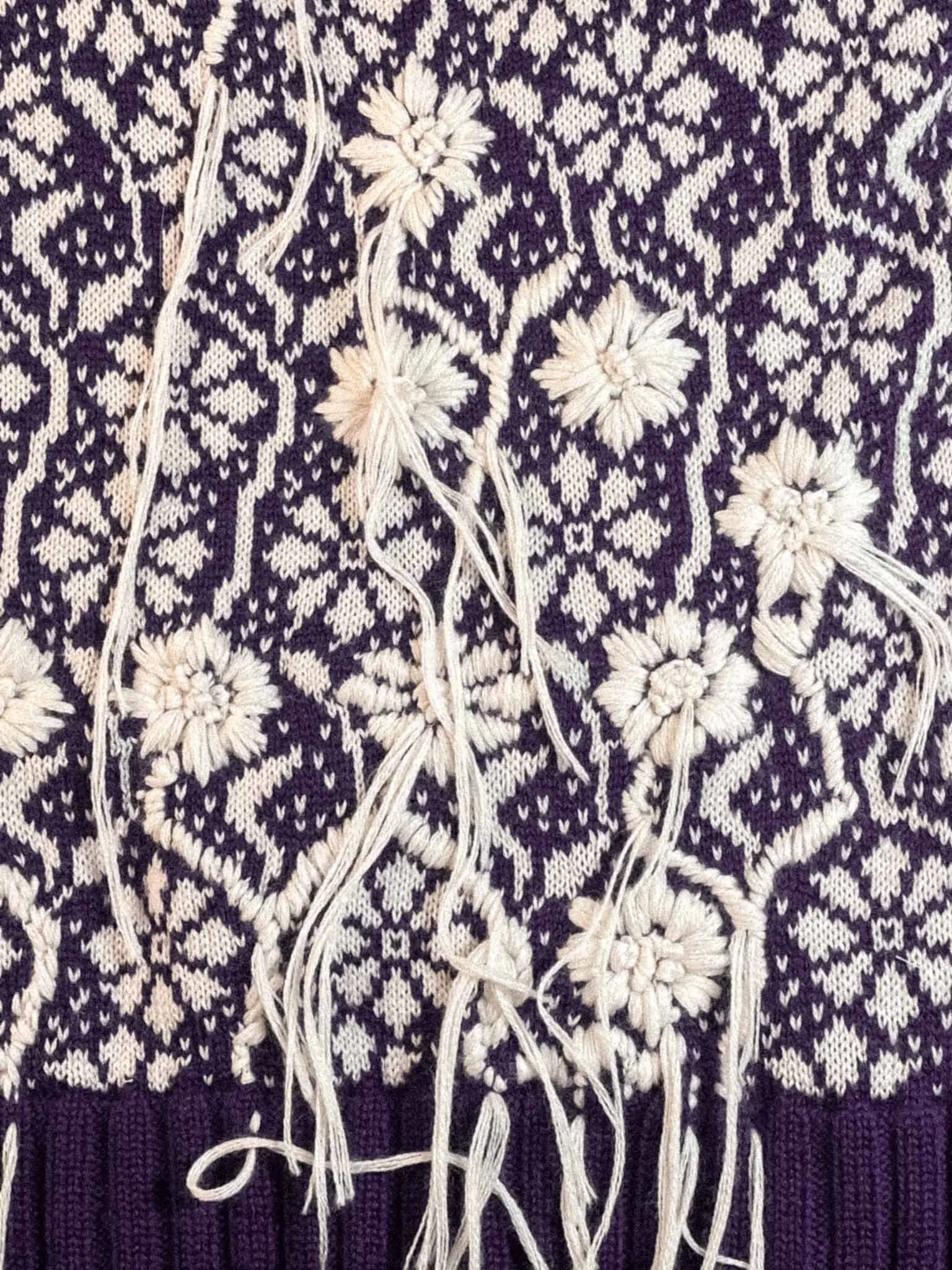 Close-up view of traditional embroidered textile with intricate white floral and geometric patterns on dark fabric. Some white thread and loose strands are visible.