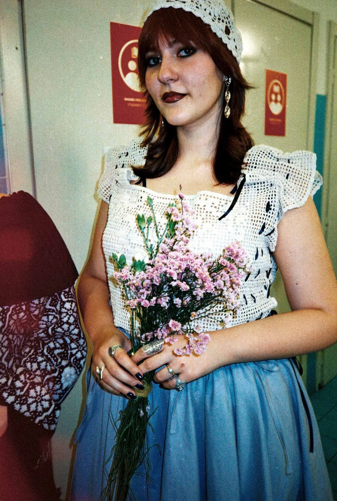 A woman with short brown hair wearing a white lace headband, black makeup, and earrings, holding a bouquet of pink flowers, dressed in a white crochet top with ruffled sleeves and a blue skirt, standing indoors with signs on the wall in the background.