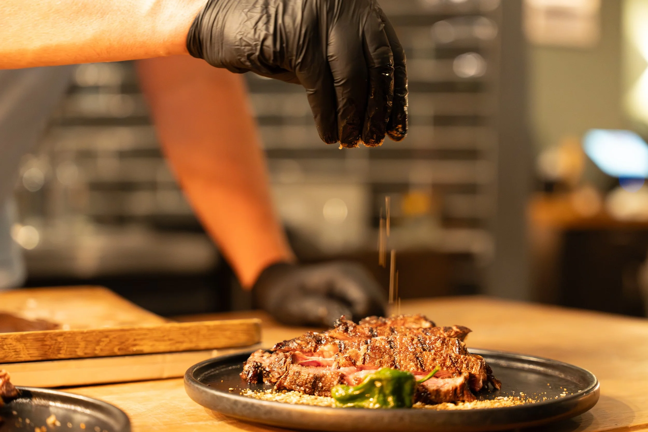 A gloved hand sprinkles seasoning onto a grilled steak on a dark plate. A small green pepper sits beside the steak on a wooden table, creating a rustic dining atmosphere.
