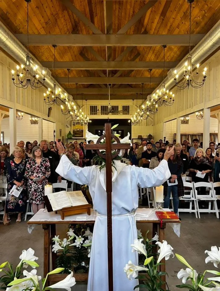 Clergy member in white robe leads a congregation during a church service, with congregation members seated in rows of chairs.